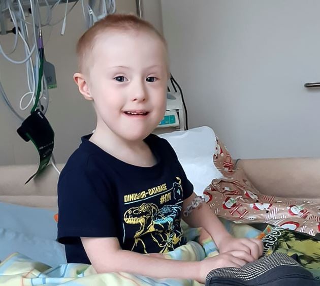 Junge mit kurzem Haar sitzt in einem Krankenhausbett und lächelt. Er trägt ein dunkles T-Shirt mit Dinosaurier-Aufdruck. Im Hintergrund sind medizinische Geräte und Schläuche zu sehen. Neben ihm liegt Geschenkpapier mit Weihnachtsmotiven.