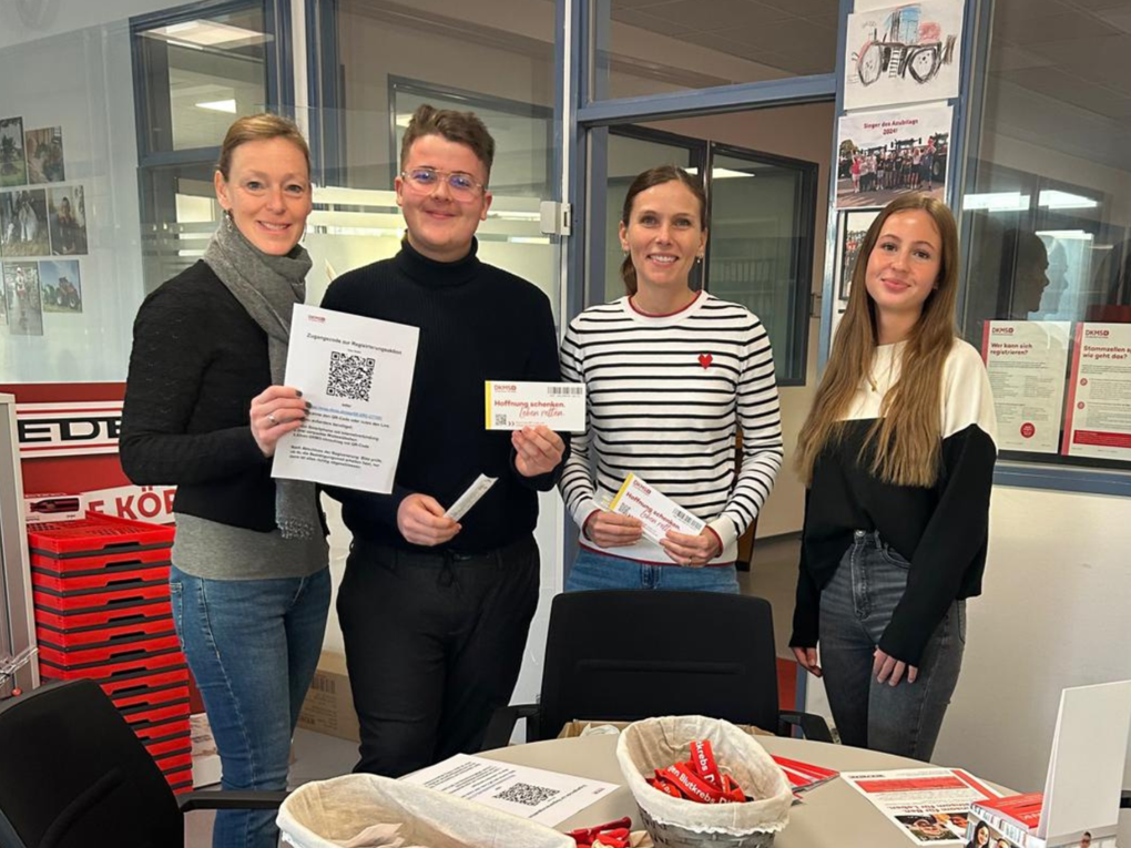 Foto von vier Mitarbeiter:innen in einem Büro, auf dem Tisch liegen DKMS-Materialien für eine Firmenaktion