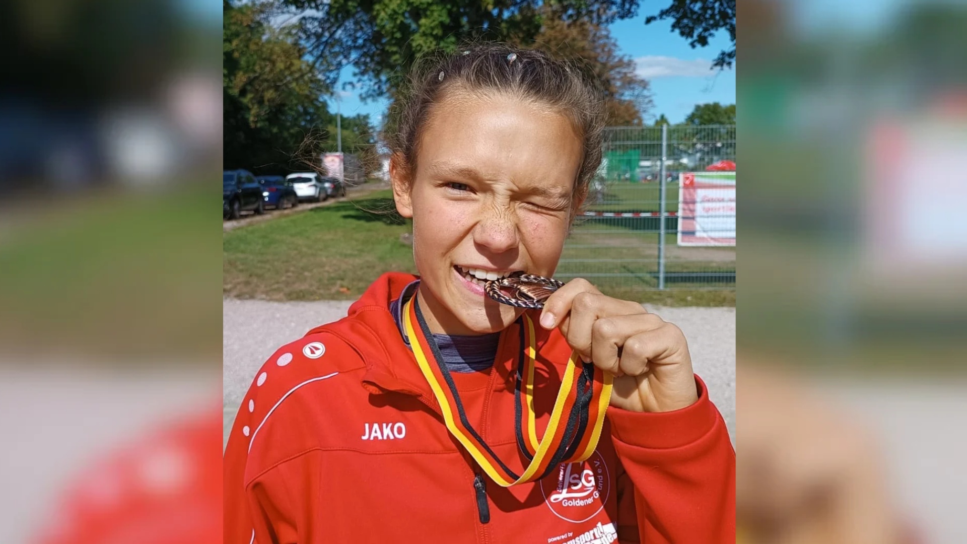 Ein Mädchen trägt eine rote Trainingsjacke und beißt auf eine Bronzemedaille.