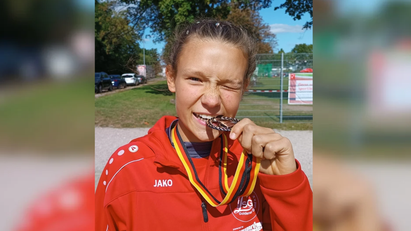 Ein Mädchen trägt eine rote Trainingsjacke und beißt auf eine Bronzemedaille.