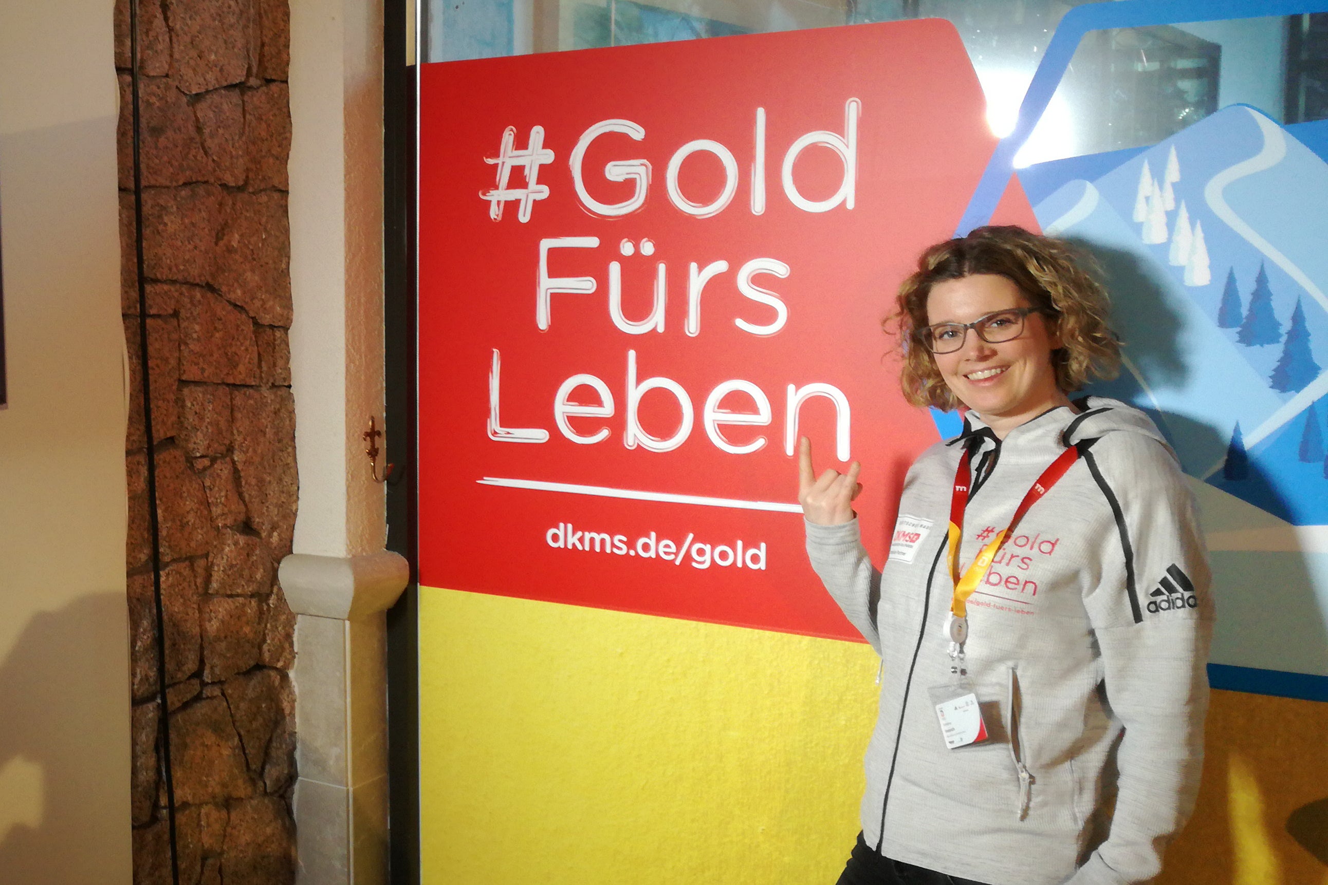 Eine Frau steht vor einem Schild der DKMS mit dem Text #Gold Fürs Leben