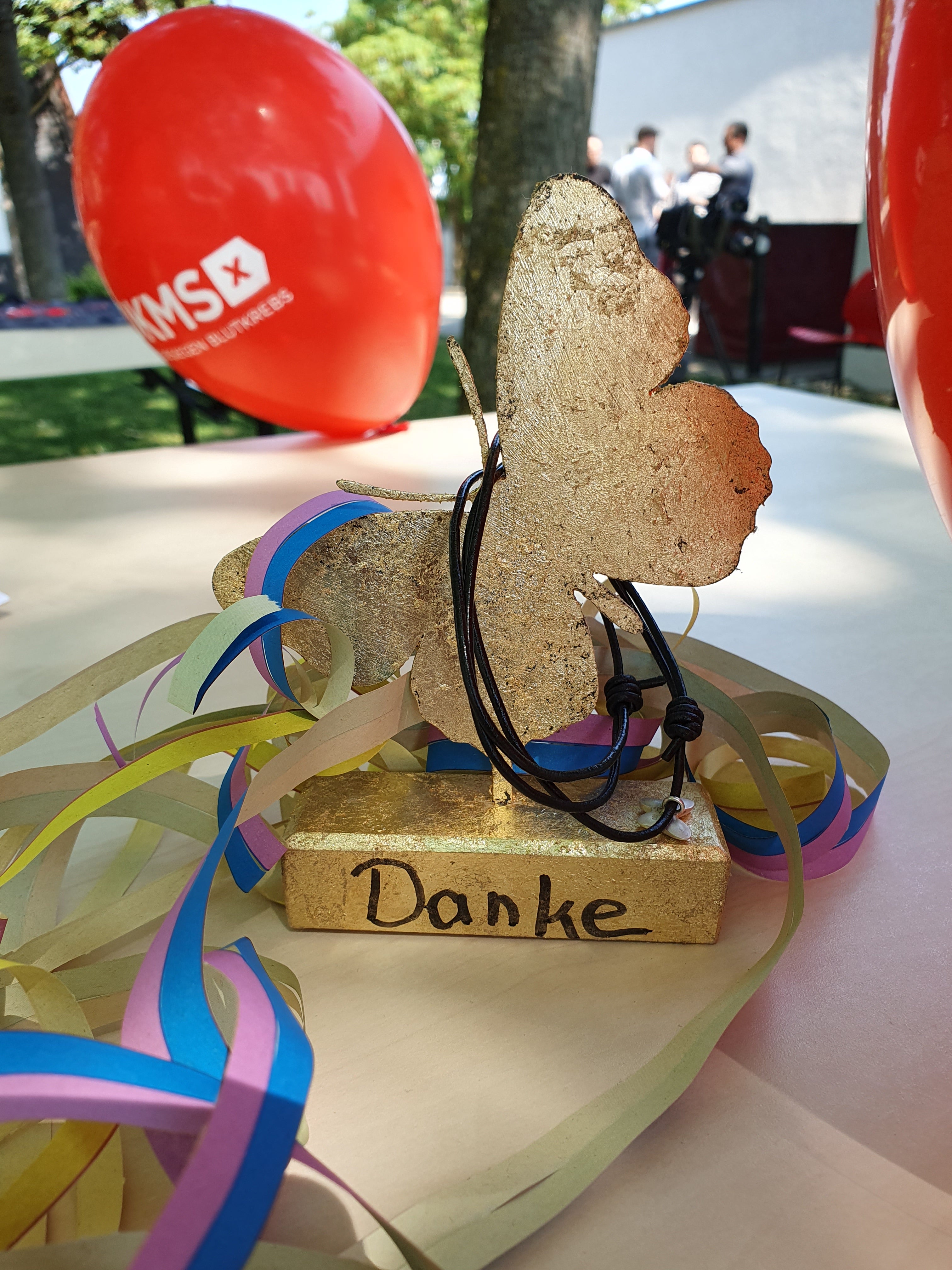 Ein goldener Schmetterling aus Holz steht auf einem Sockel mit der Aufschrift „Danke“. Um das Objekt sind bunte Papierbänder und ein schwarzes Band gelegt. Im Hintergrund sind rote DKMS-Ballons, ein Baum und mehrere Personen auf einer Wiese zu sehen.
