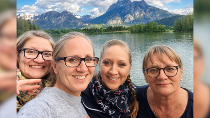 Vier Frauen stehen lächelnd vor einem Gewässer, am Horizont ist ein großer Berg zu sehen. 
