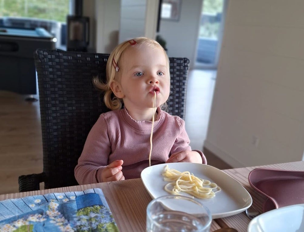 Ein kleines blondes Mädchen sitzt an einem Tisch. Vor ihr steht ein Teller mit Spaghetti und sie hat eine im Mund.