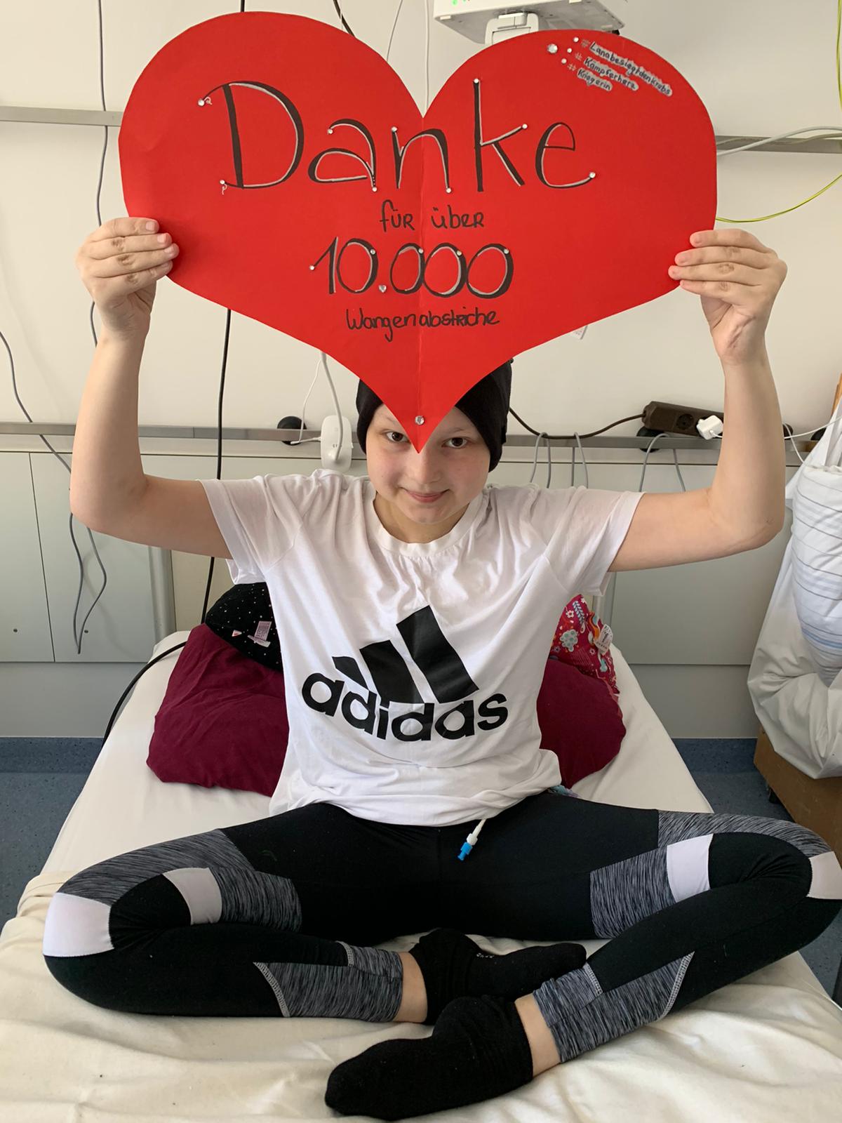 Lana sitzt im Schneidersitz auf einem Bett und hält ein Schild: "Danke für über 10 Tausend Wangenabstriche"