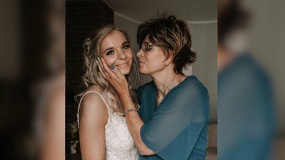 Eine ältere Frau trägt ein grünblaues Kleid und hält das Gesicht einer jungen, ein Hochzeitskleid tragenden Frau in den Händen. 