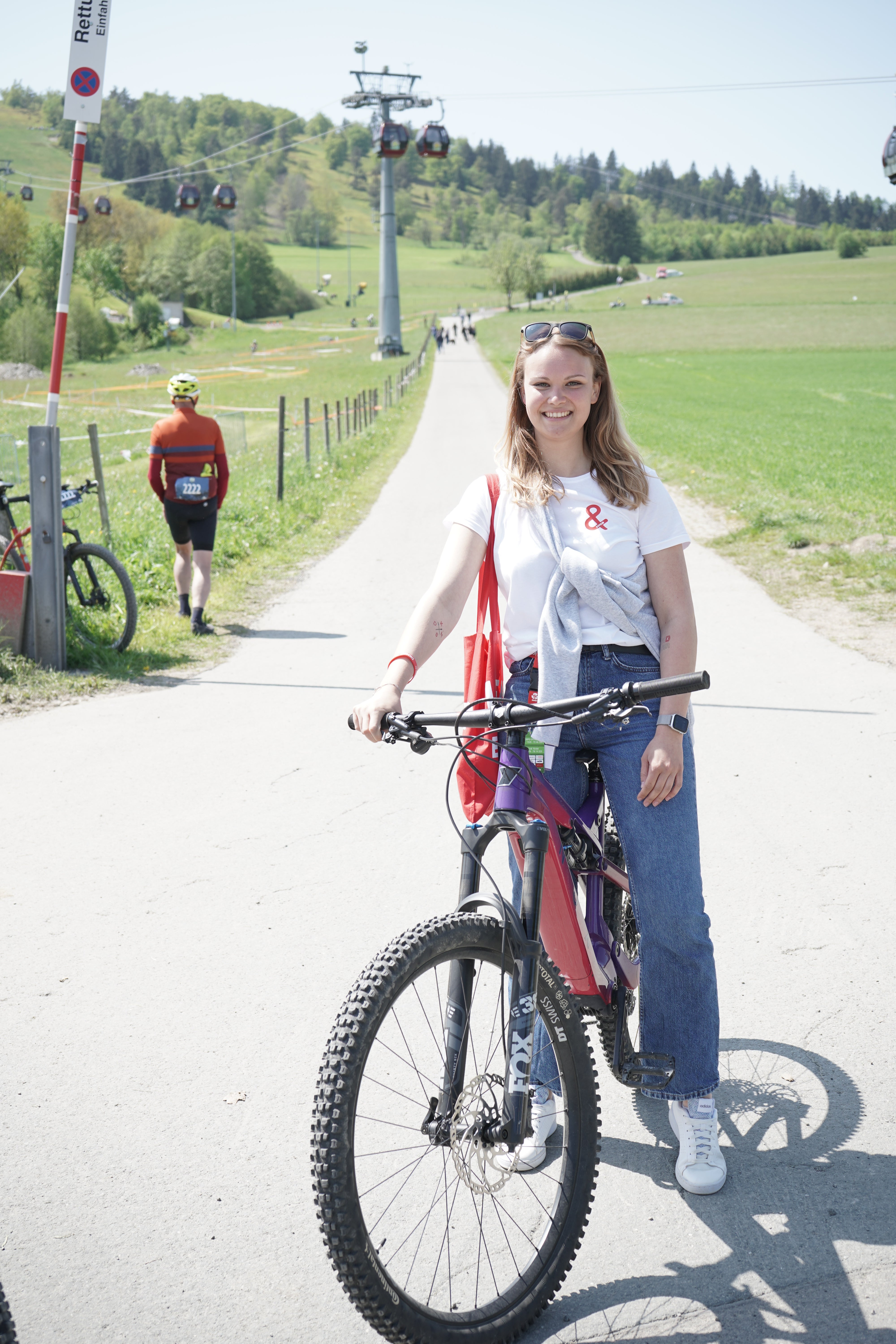 Eine Frau steht mit einem Mountainbike auf einem asphaltierten Weg in einer ländlichen Umgebung. Sie trägt ein weißes T-Shirt mit rotem &-Symbol, blaue Jeans und weiße Schuhe. Im Hintergrund sind Wiesen, ein Skilift mit Gondeln, ein Radfahrer in rotem Trikot und mehrere Personen zu sehen.