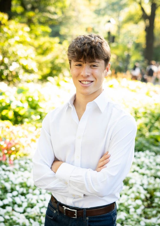 Ein Junge steht mit verkreuzten Armen und lächelt. Er hat braune Haare und trägt ein weißes Hemd.