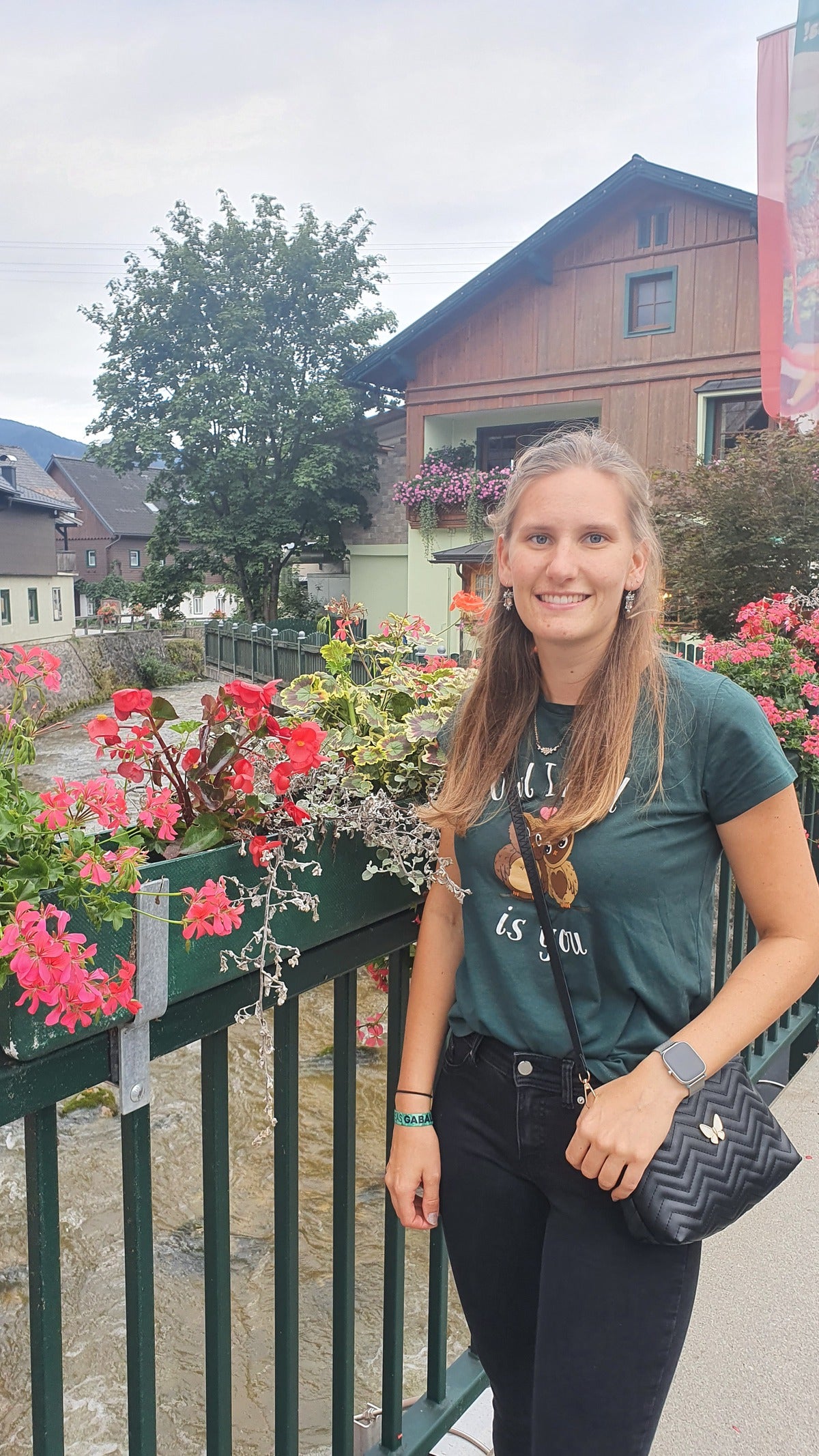 Eine junge Frau steht lächelnd an einem grünen Geländer, das mit pinken und roten Blumen geschmückt ist. Im Hintergrund sind ein kleiner Fluss, ein Haus mit Blumenkästen am Balkon und ein Baum zu sehen. Die Frau trägt ein dunkelgrünes T-Shirt, eine schwarze Hose und eine schwarze Umhängetasche. Es ist bewölkt.