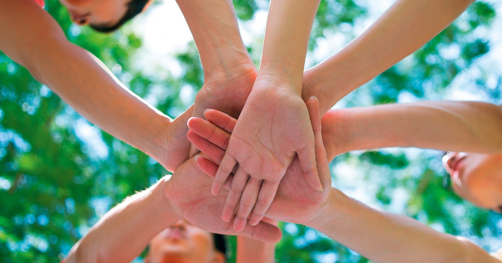 Mehrere Menschen legen im Kreis stehend ihre Hände übereinander, als Zeichen von Teamgeist und Zusammenhalt. Der Hintergrund zeigt grünes Laub und blauen Himmel, die Perspektive ist von unten aufgenommen.