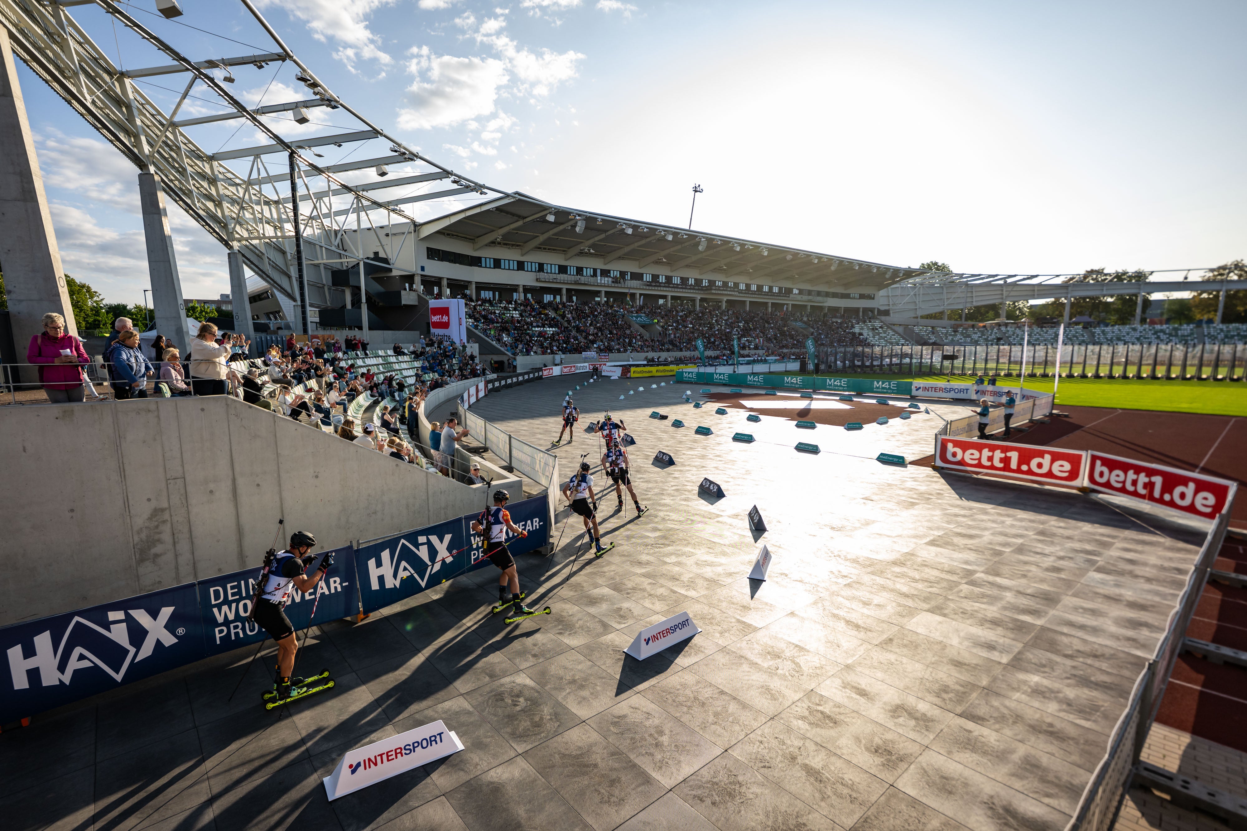 Sonnenuntergang über der Rennstrecke im Heinz-Speyer-Stadion in Dresden. Biathleten fahren an den Rängen vorbei. 