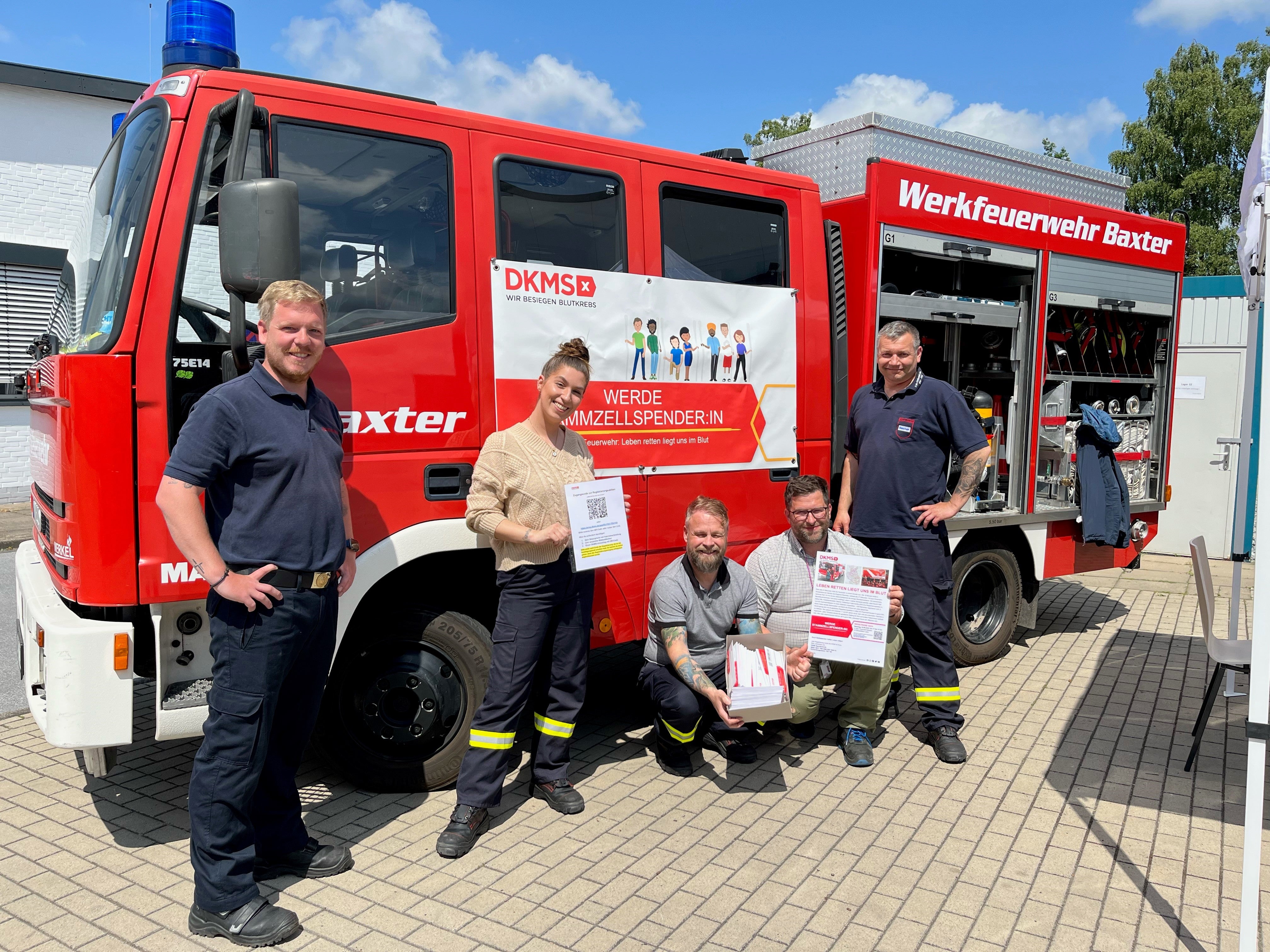 Fünf Personen stehen oder knien vor einem roten Einsatzfahrzeug der Werkfeuerwehr Baxter. Zwei Personen halten DKMS-Infomaterial und eine Box mit Registrierungssets. Am Fahrzeug ist ein DKMS-Banner mit der Aufschrift „Werde Stammzellspender\:in!“ angebracht. Die Seitenfächer des Fahrzeugs sind geöffnet und zeigen Feuerwehr-Ausrüstung. Der Boden ist gepflastert, im Hintergrund sind ein Gebäude und Bäume zu sehen.

