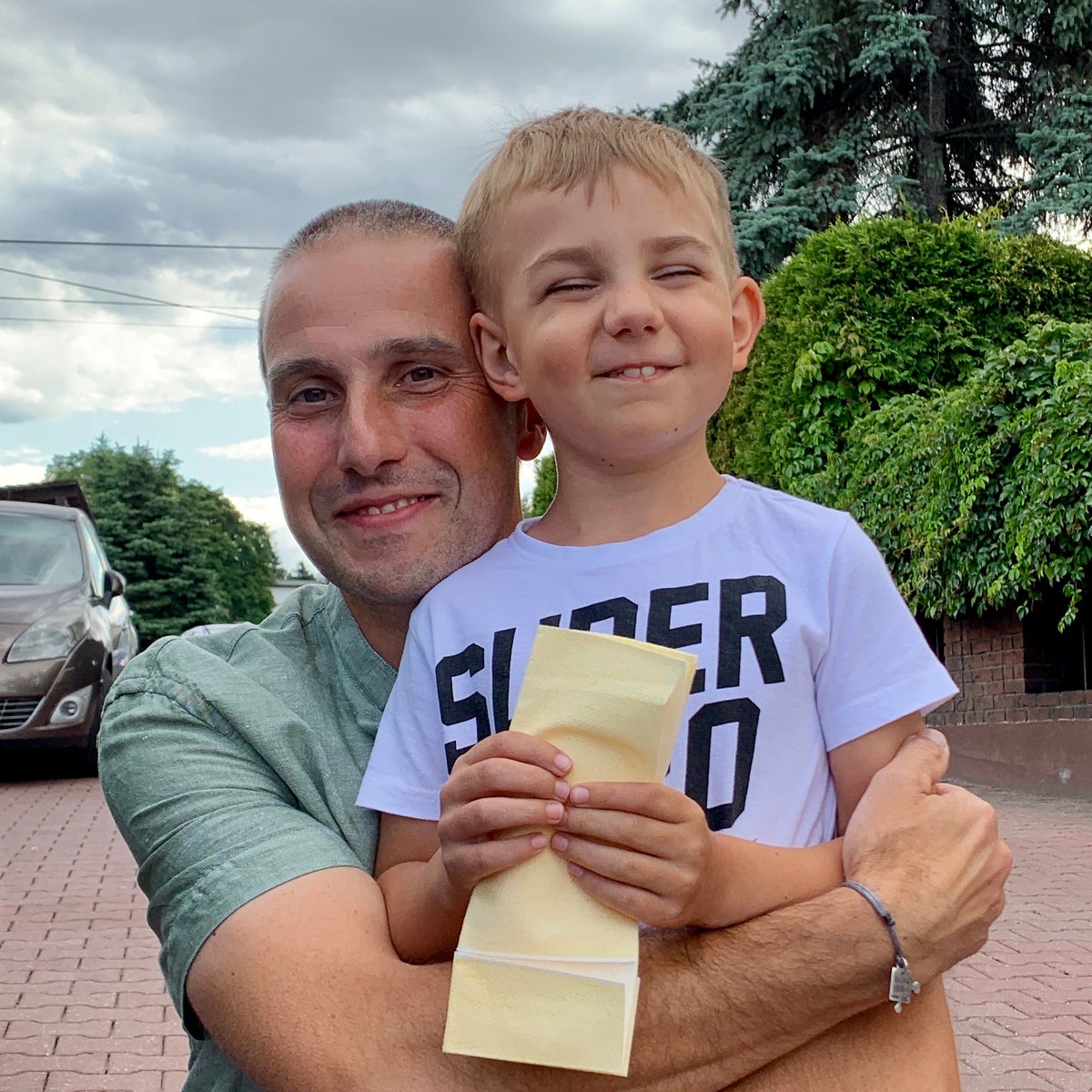 Ein Mann hält ein Kind im Arm, beide lächeln in die Kamera. Der Junge trägt ein weißes T-Shirt mit schwarzer Aufschrift und hält einen gelben Umschlag in den Händen. Der Mann trägt ein grünes T-Shirt. Im Hintergrund sind ein gepflasterter Hof, ein Auto, grüne Sträucher und Bäume sowie ein bewölkter Himmel zu sehen.