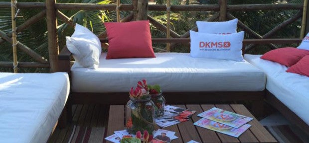 Eine weiße Couch mit Kissen, auf zwei Kissen sind DKMS Logos gedrückt. Davor ein Tisch mit Blumen und Zeitschriften