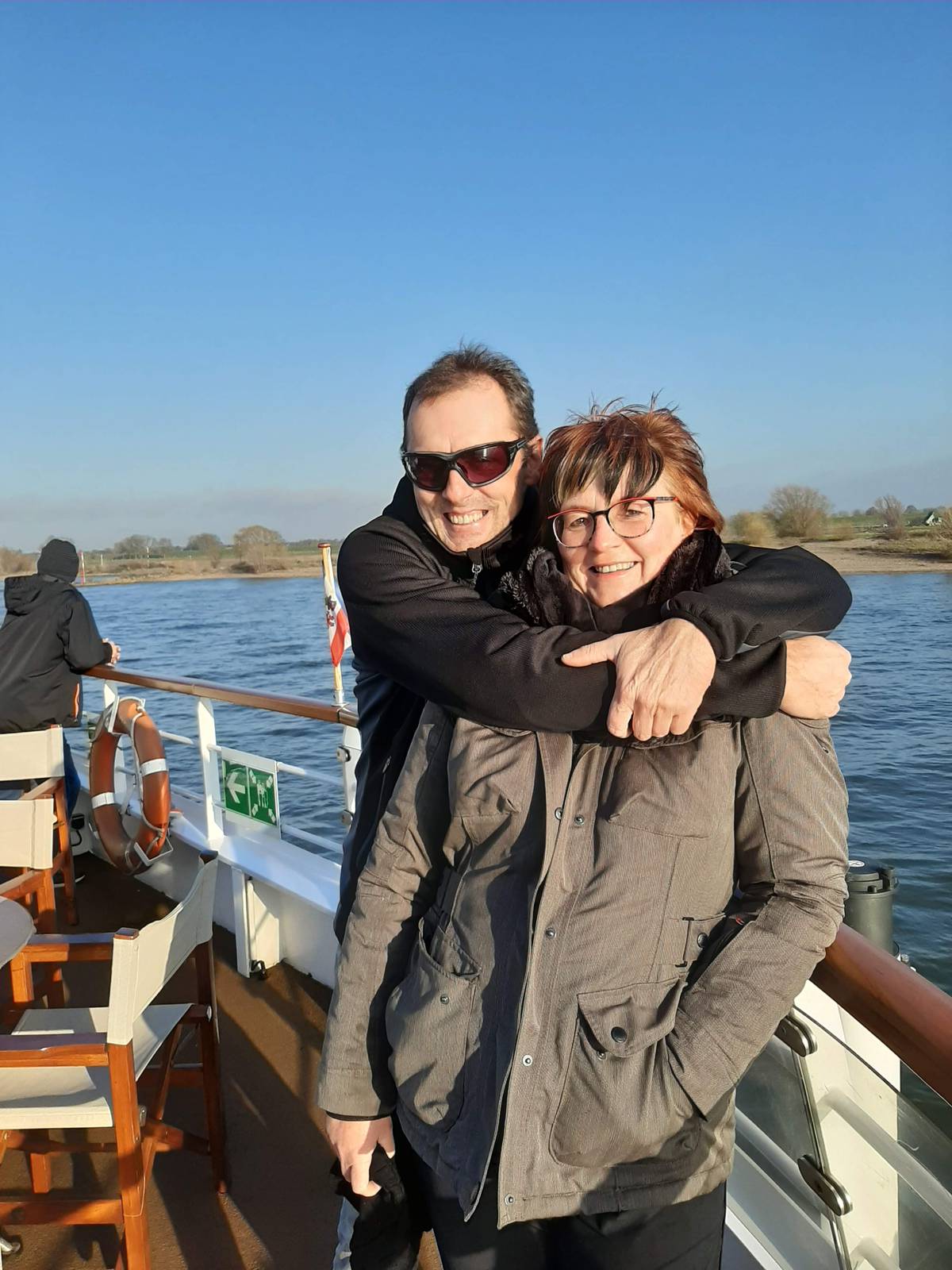 Ein Mann steht auf einem Schiff hinter einer Frau und umarmt sie von hinten. Beide tragen Jacken, der Mann eine Sonnenbrille. Sie stehen an der Reling, im Hintergrund ist Wasser, Ufer mit Bäumen und blauer Himmel zu sehen. Auf dem Deck stehen mehrere Stühle.