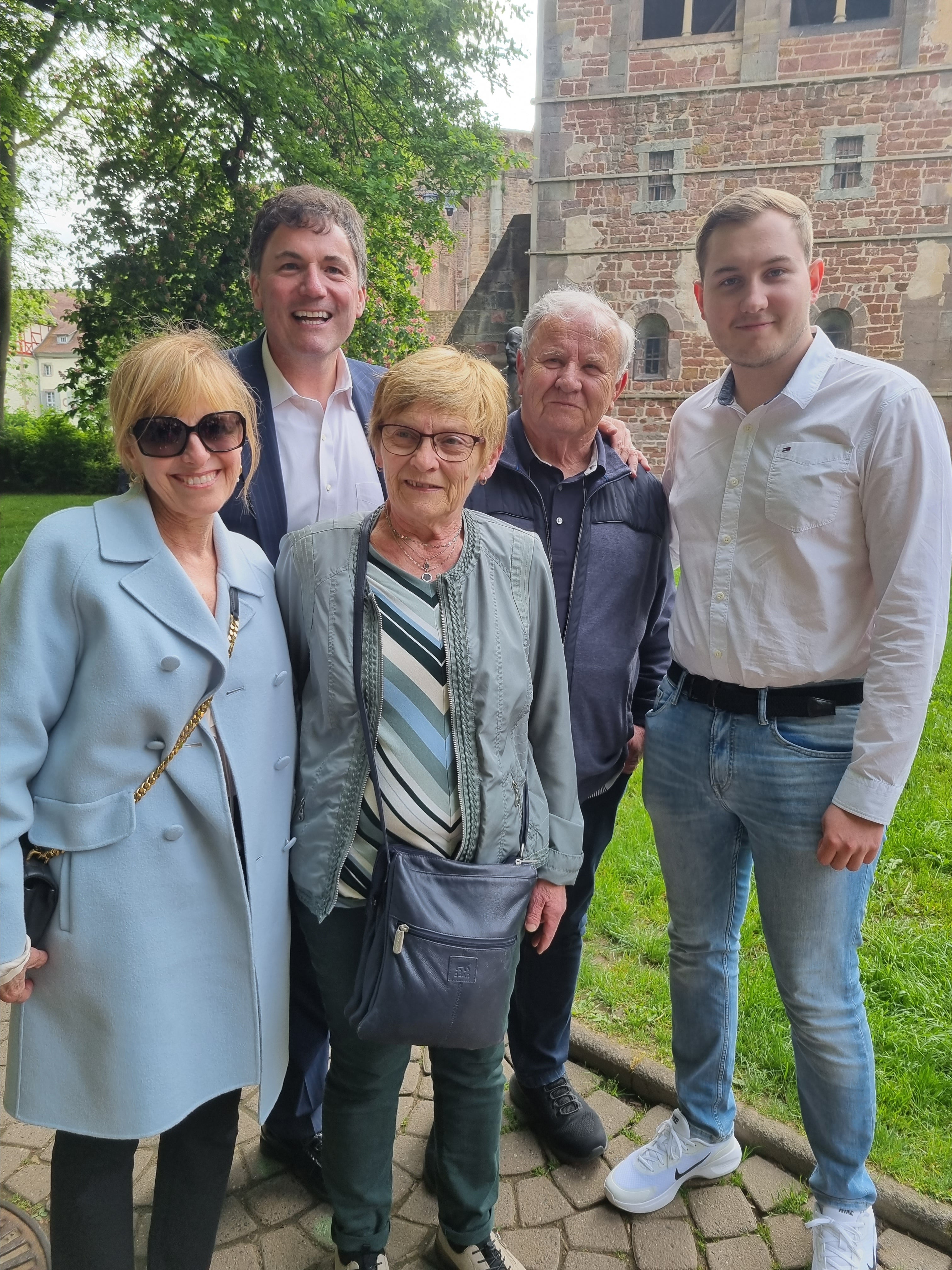 Fünf Personen stehen auf einem gepflasterten Weg vor einer Rasenfläche und einem Gebäude mit Backsteinfassade. Zwei Männer und drei Frauen blicken in die Kamera. Die Personen tragen Jacken, Hemden oder Blusen. Im Hintergrund sind Bäume und ein Teil eines historischen Gebäudes zu sehen.