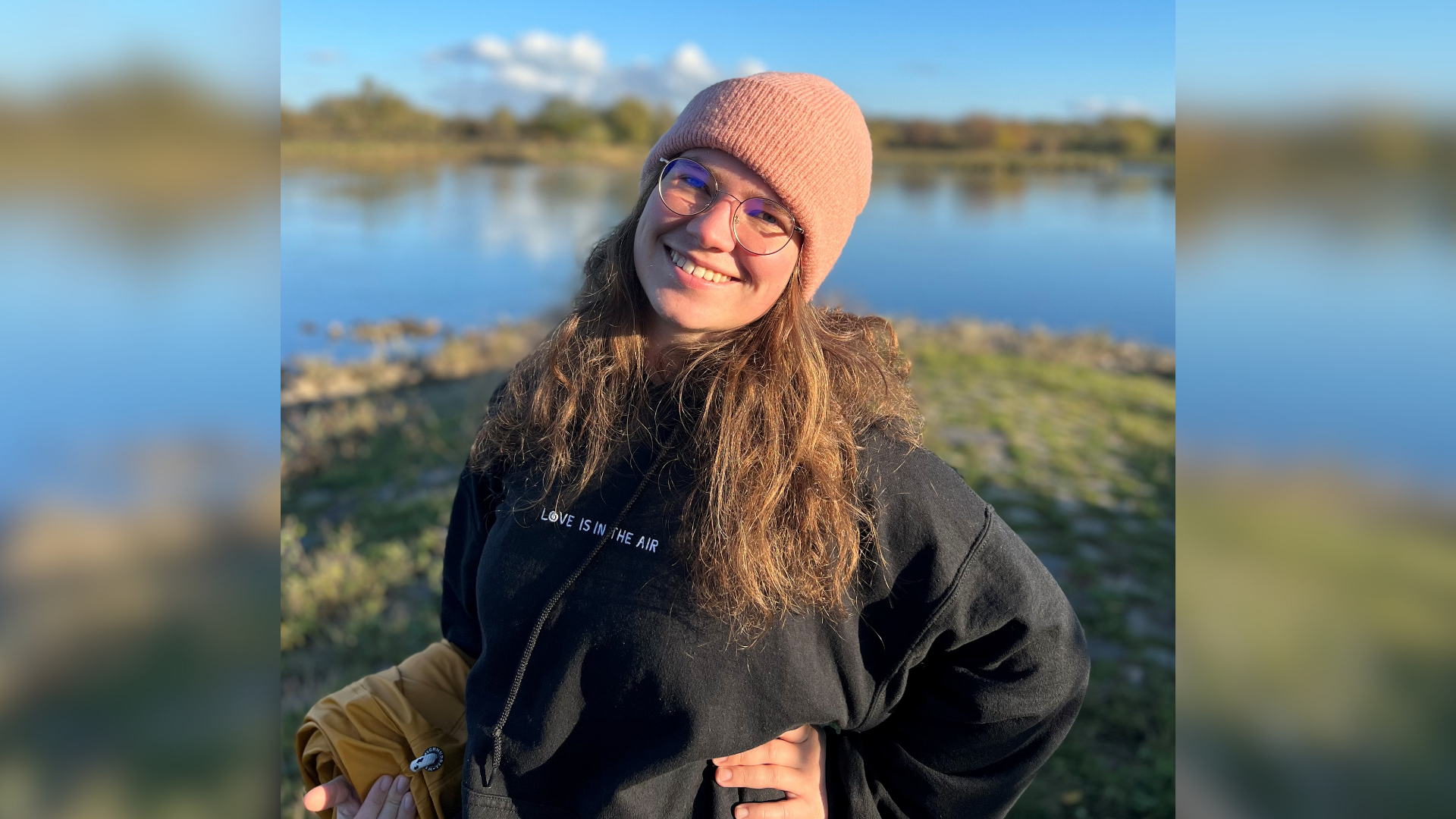 Eine Frau trägt eine rosafarbene Mütze und einen schwarzen Kapuzenpullover. Sie steht am Ufer eines Gewässers. 