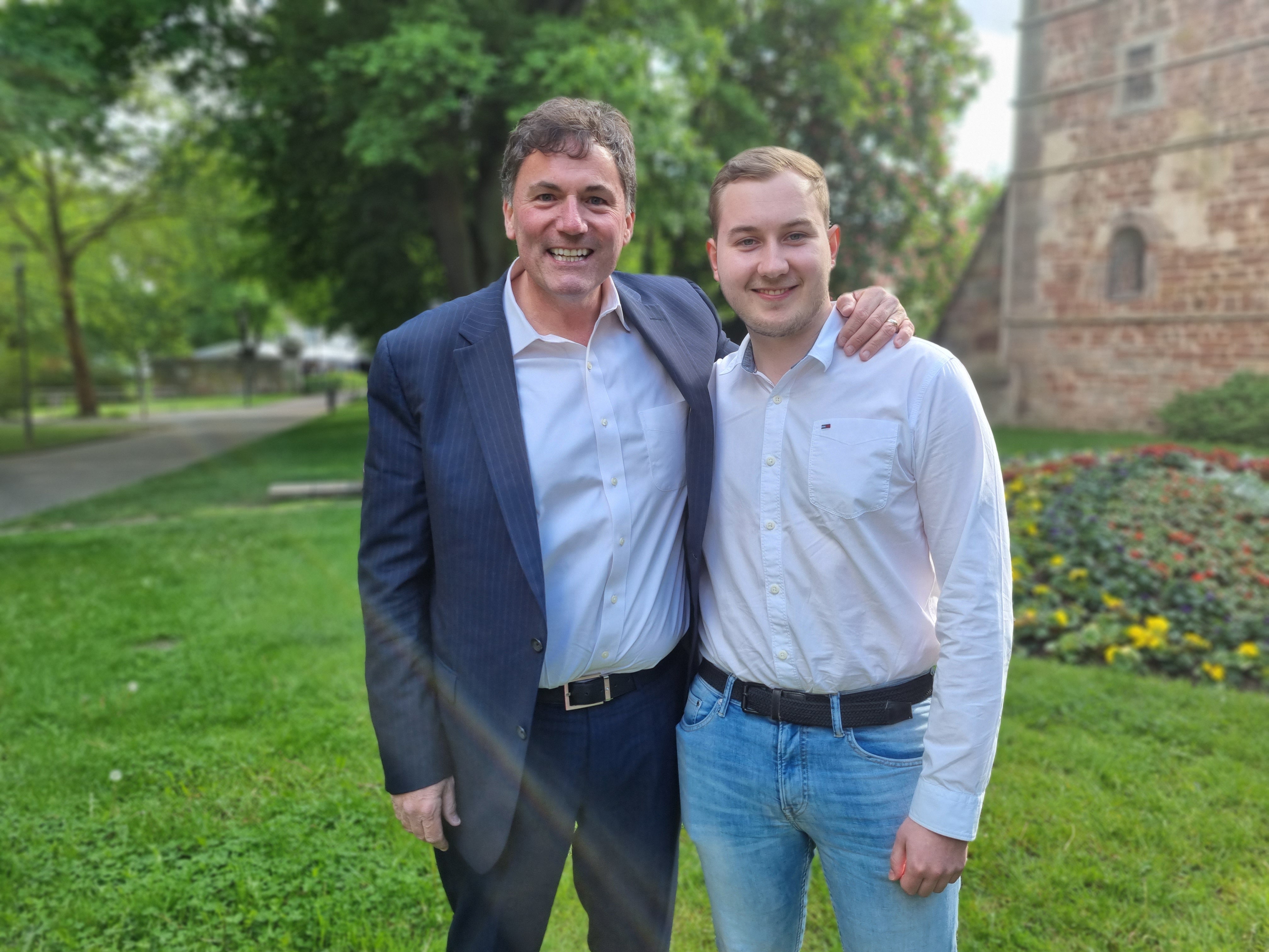 Zwei Männer stehen nebeneinander auf einer Rasenfläche. Der Mann links trägt einen dunklen Anzug, der Mann rechts ein weißes Hemd und eine blaue Jeans. Der linke Mann hat seinen Arm um die Schulter des rechten Mannes gelegt. Im Hintergrund sind ein Blumenbeet, Bäume, ein Weg und ein Gebäude mit Backsteinfassade zu sehen.