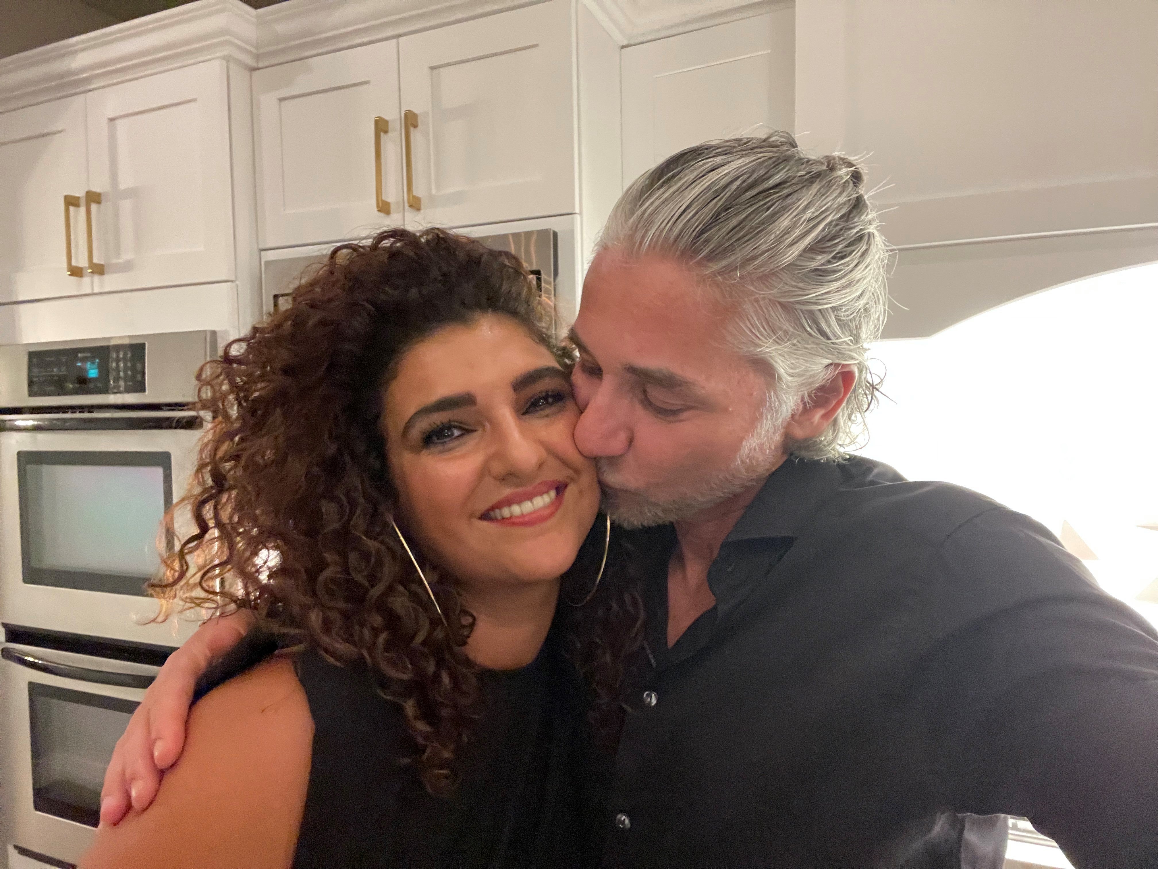 Ein Mann küsst eine Frau auf die Wange. Im Hintergrund sieht man eine Küche