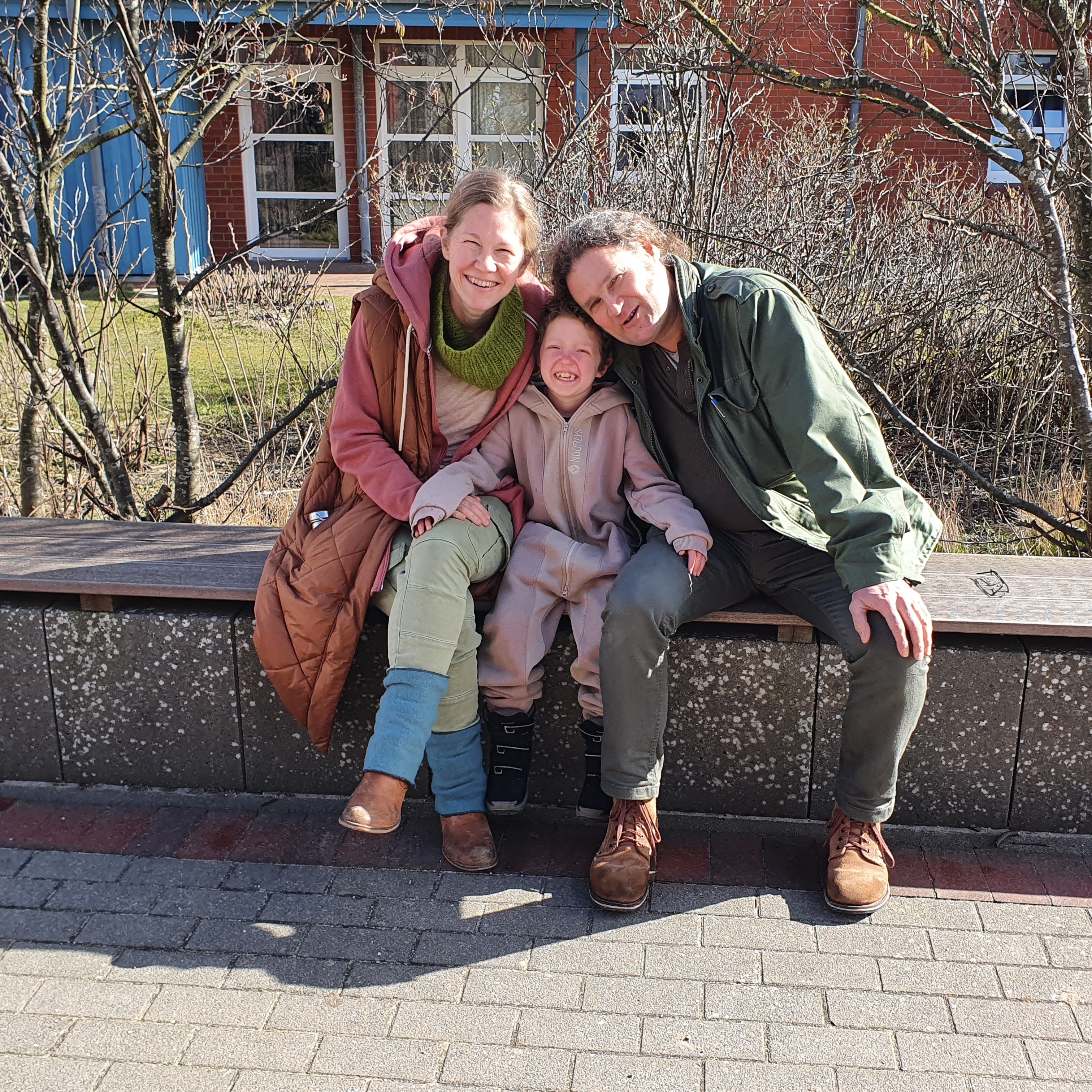 Ein fröhliches Familienfoto im Freien. Zwei Erwachsene und ein Kind sitzen eng aneinandergeschmiegt auf einer Holzbank und lächeln in die Kamera. Die Erwachsene links trägt eine orangefarbene Jacke und einen grünen Schal, das Kind in der Mitte trägt einen beigefarbenen Einteiler, der Erwachsene rechts eine grüne Jacke. Im Hintergrund sind kahle Büsche, ein rotes Backsteingebäude und Fenster mit weißen Rahmen zu sehen. 
