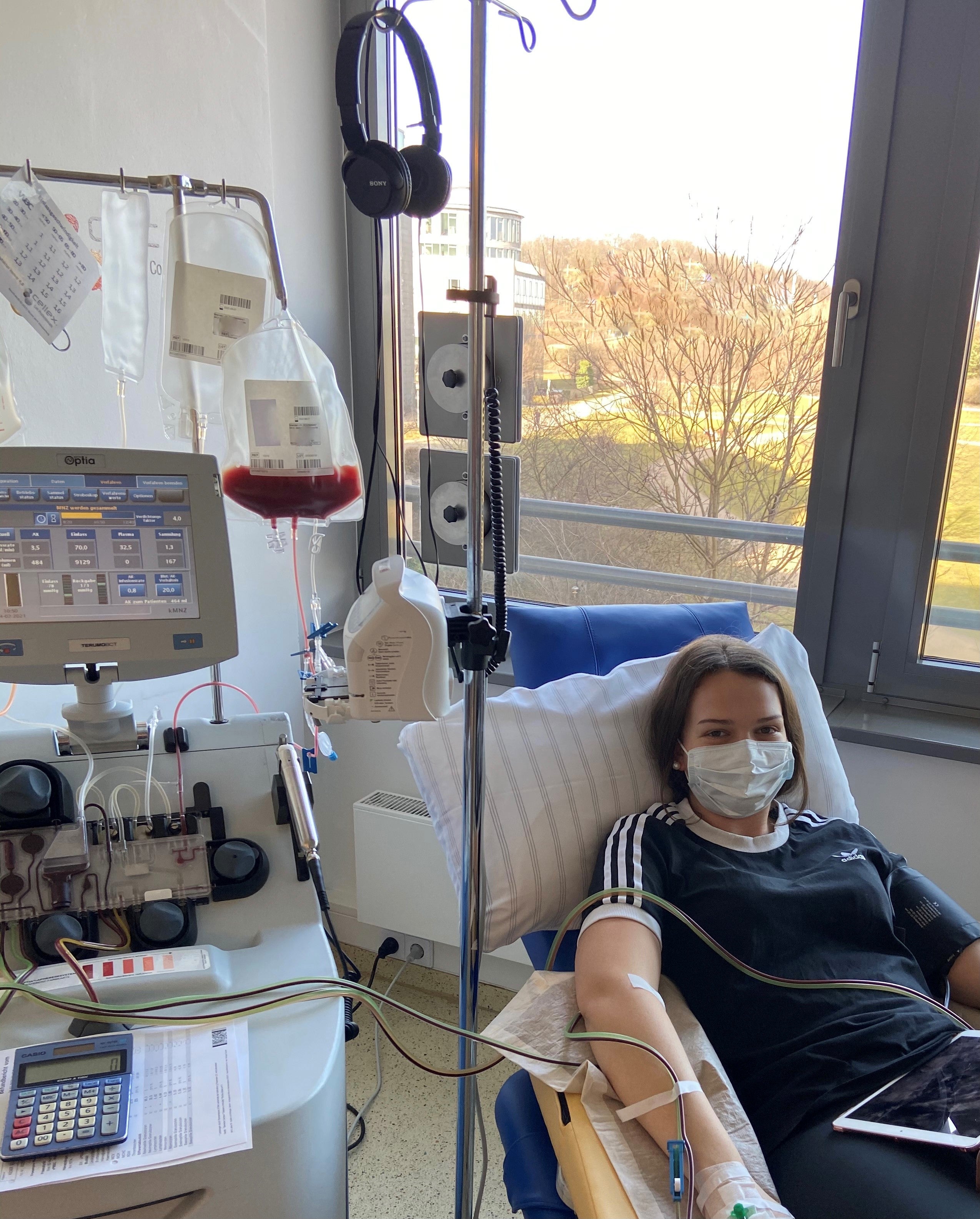 Eine Frau liegt in einem medizinischen Behandlungsstuhl und spendet periphere Blutstammzellen. Sie trägt eine medizinische Maske und ein dunkles T-Shirt. Ihr rechter Arm ist an eine Apheresemaschine angeschlossen. Neben ihr stehen Infusionsbeutel, ein Monitor und medizinisches Zubehör. Durch das Fenster ist eine Landschaft mit Bäumen und Wiese zu sehen.