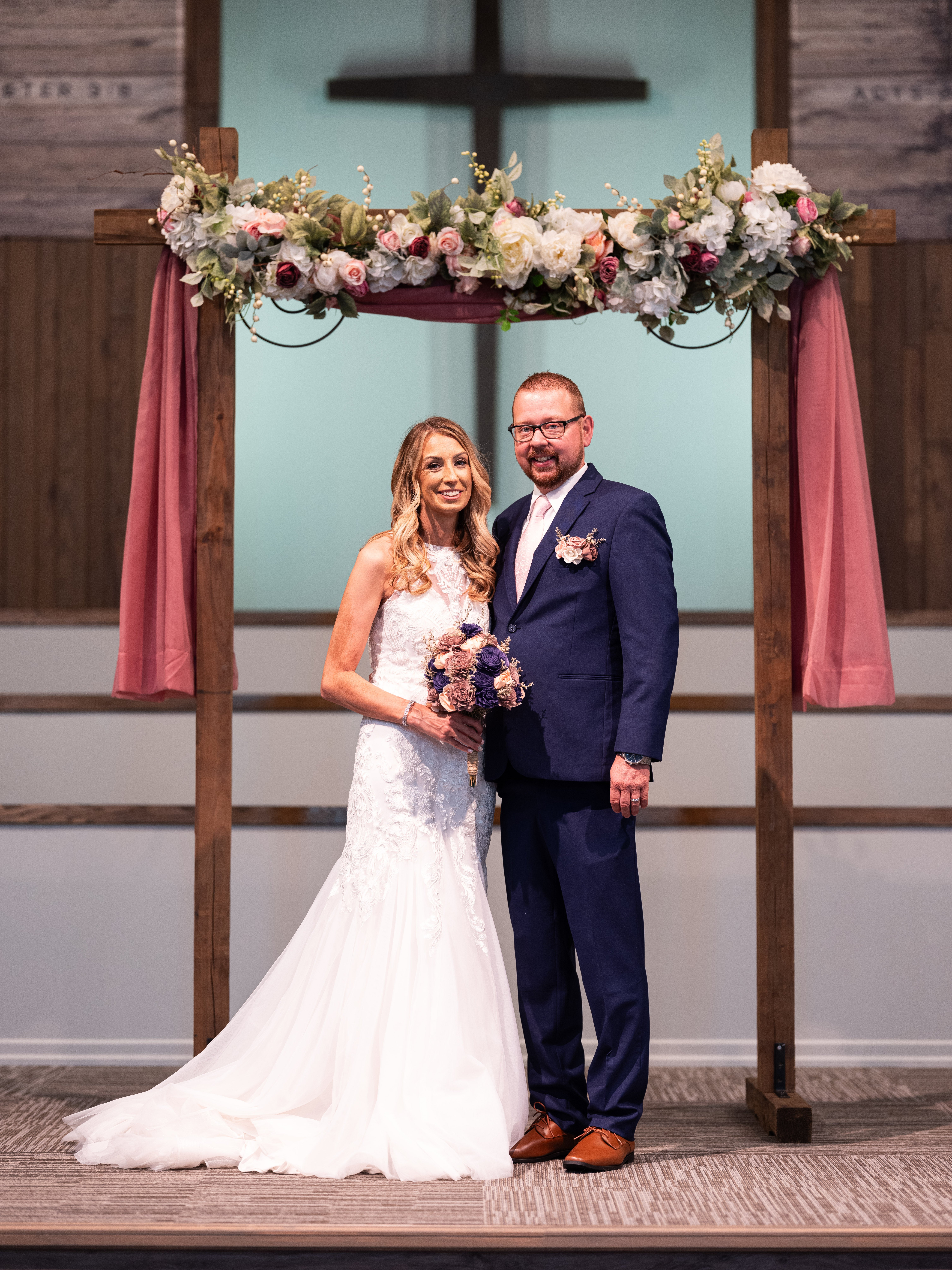 Ein Brautpaar steht unter einem blumengeschmückten Holzbogen in einem festlich dekorierten Innenraum. Die Braut trägt ein weißes, bodenlanges Kleid mit Spitze und hält einen Blumenstrauß in der Hand. Der Bräutigam trägt einen dunkelblauen Anzug mit Weste und Ansteckblume. Im Hintergrund ist ein Kreuz zu sehen.