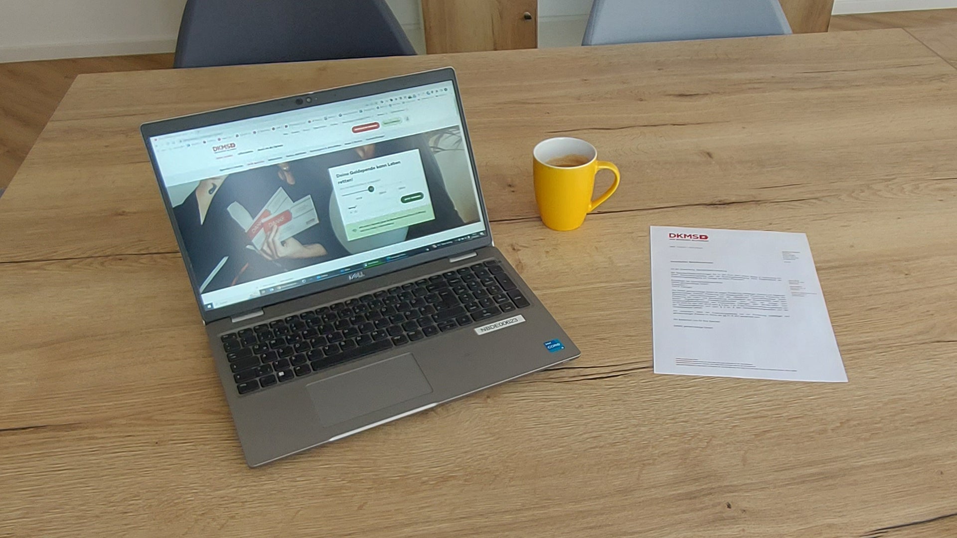 Holztisch mit einem geöffneten Laptop, auf dessen Bildschirm eine Spendenseite der DKMS zu sehen ist. Daneben stehen eine gelbe Tasse und ein ausgedrucktes DKMS-Dokument. Im Hintergrund sind zwei Stühle sichtbar.