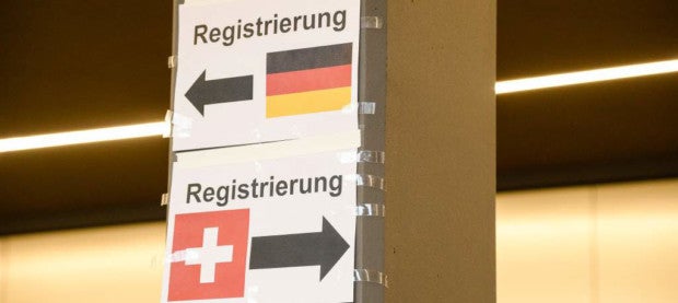 Zwei Schilder mit der Deutschen und Schweizer Flagge, beide mit dem Aufdruck Registrierung und Pfeilen die jeweils in unterschiedliche Richtungen zeigen