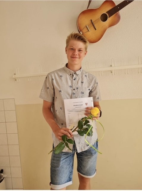Ein Jugendlicher steht in einem Raum vor einer hellen Wand mit Hakenleiste und hält ein Dokument sowie eine gelbe Rose in der Hand. Er trägt ein kurzärmliges Hemd mit Palmenmuster und eine kurze Jeanshose. Über ihm hängt eine Akustikgitarre an der Wand.