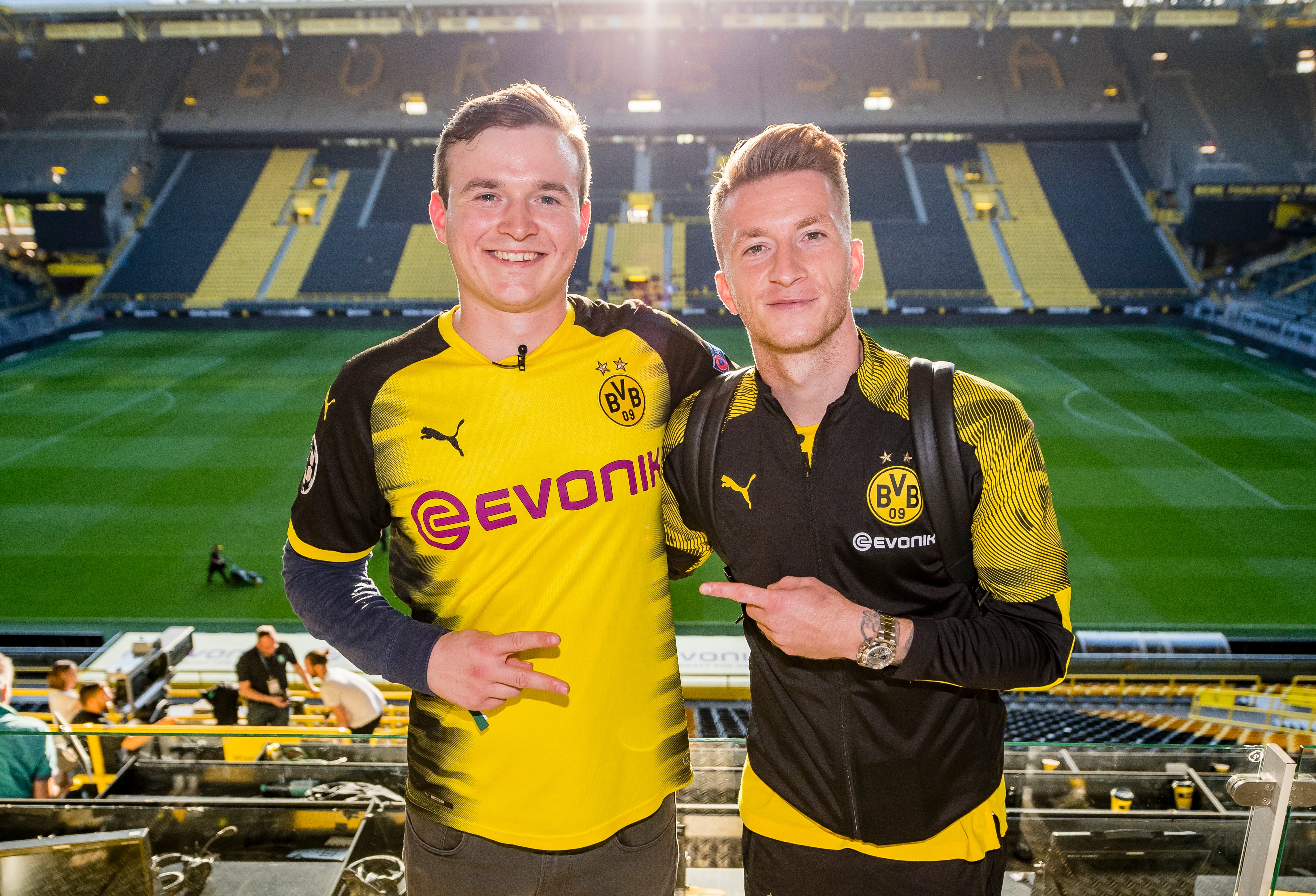 Ludwig Jolmes und Marco Reus im Stadion, beide tragen BVB Merch und im Hintergrund sieht man das BVB Stadion