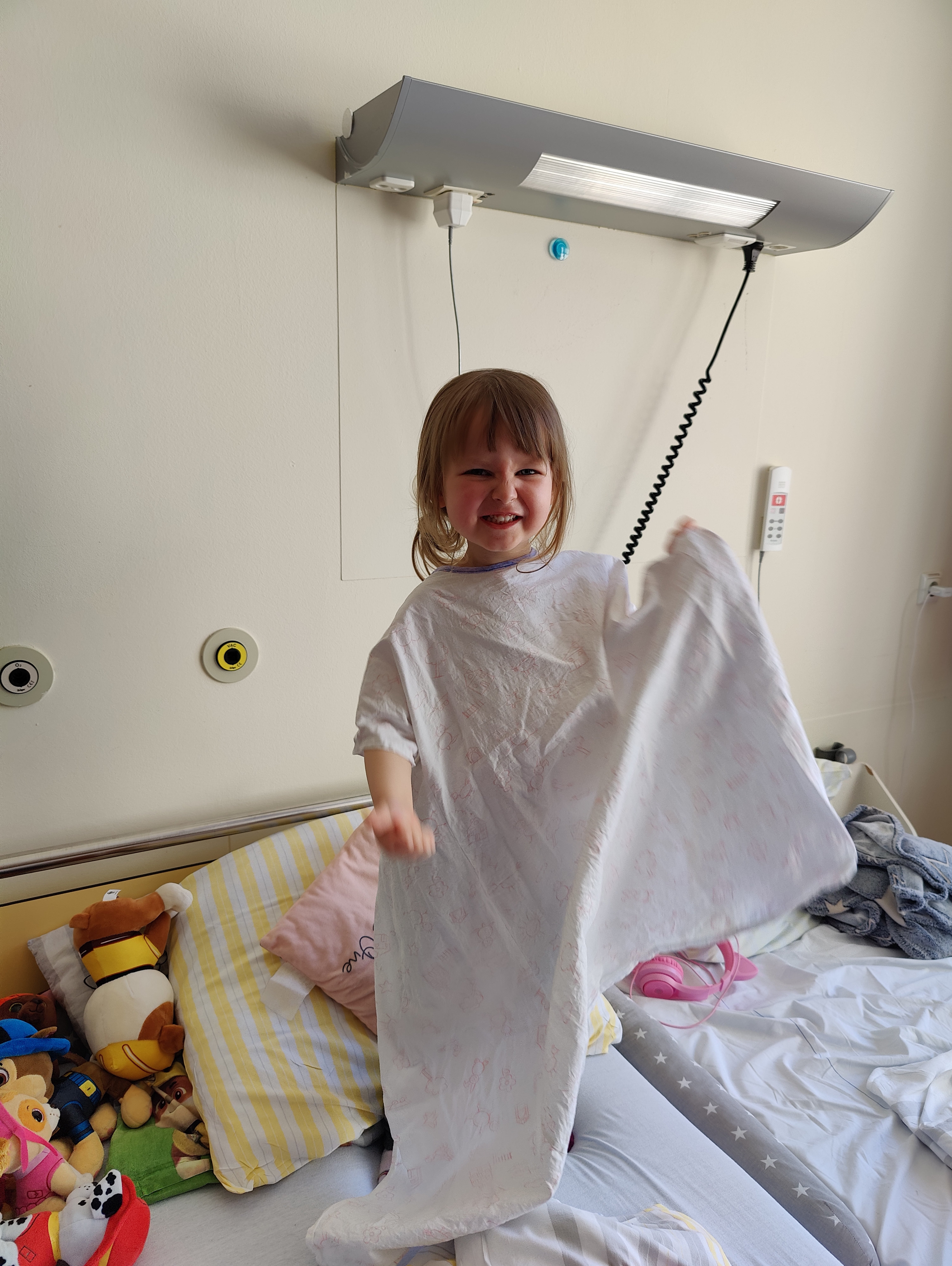 Ein kleines Kind steht lachend in einem Krankenhausbett und hält ein Tuch oder eine Decke in der Hand. Auf dem Bett liegen Kuscheltiere und Kissen.