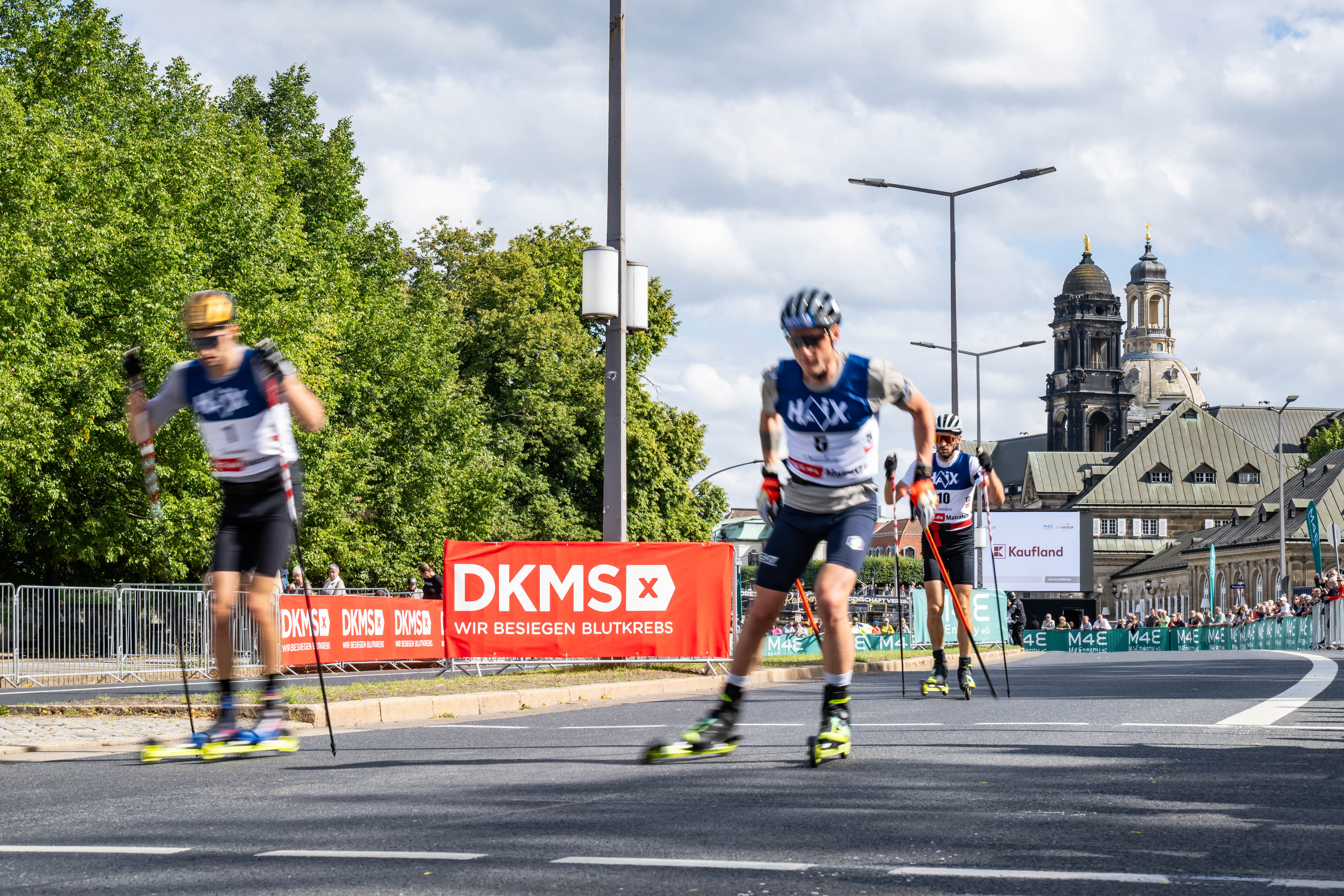 Biathlon-Athleten auf rollenden Skiern vor einem DKMS Banner und der Dresdner Skyline. 