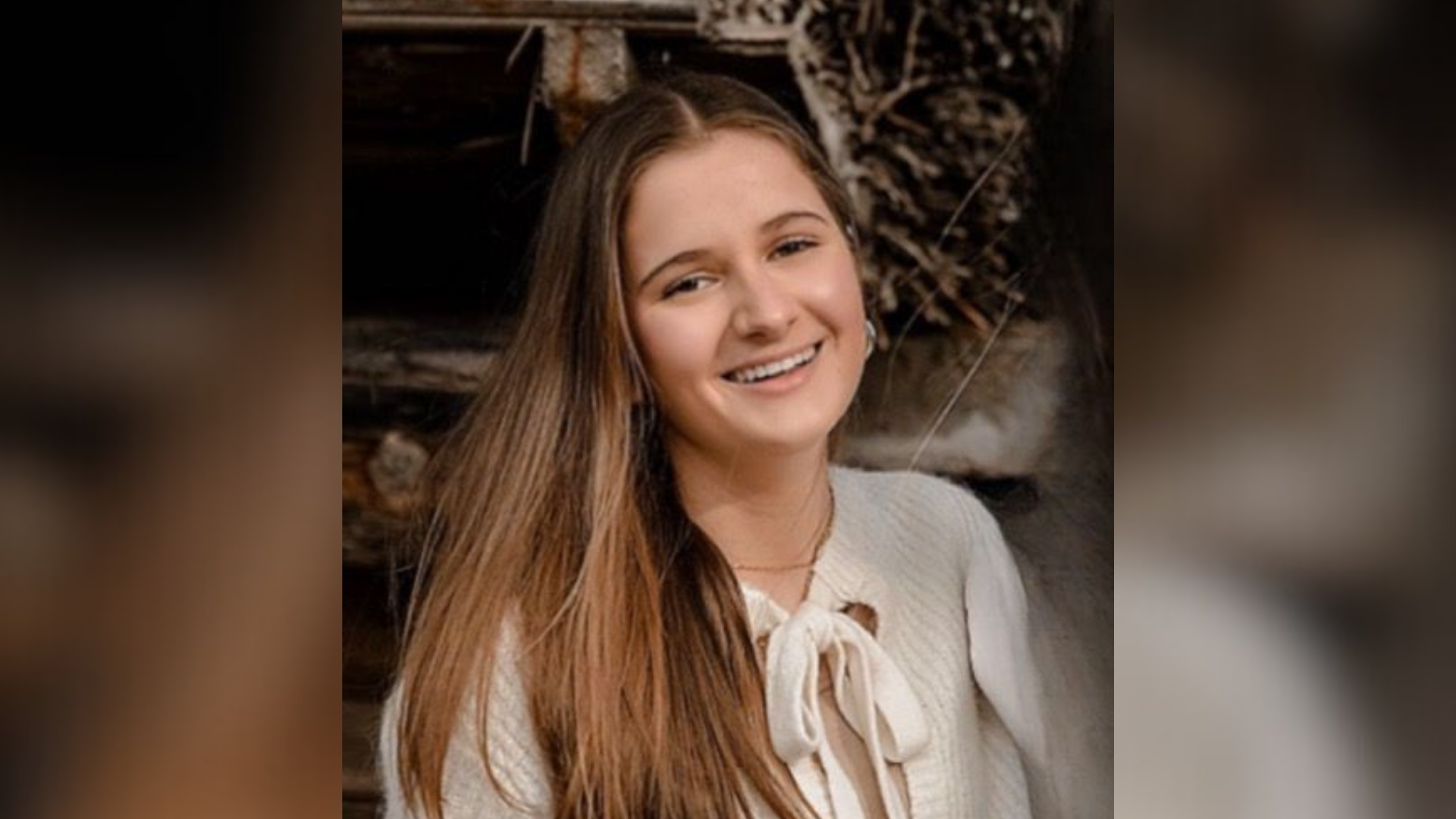 Eine junge lächelnde Frau steht vor einem Kaminholzunterstand und trägt einen weißen Strickpullover.