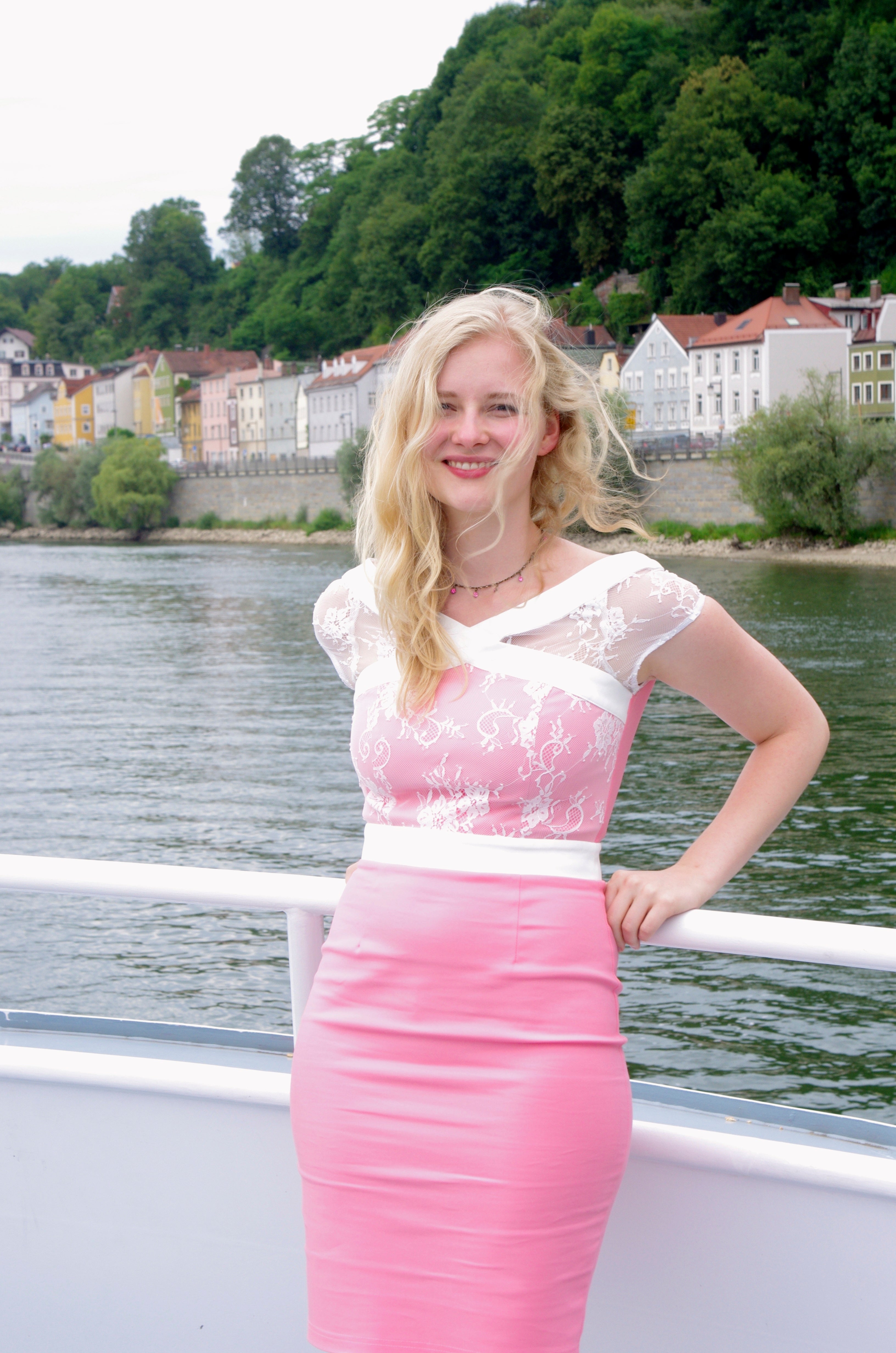 Eine Frau in einem rosa Kleid lehnt an einem Geländer auf einem Schiff, im Hintergrund ist Wasser