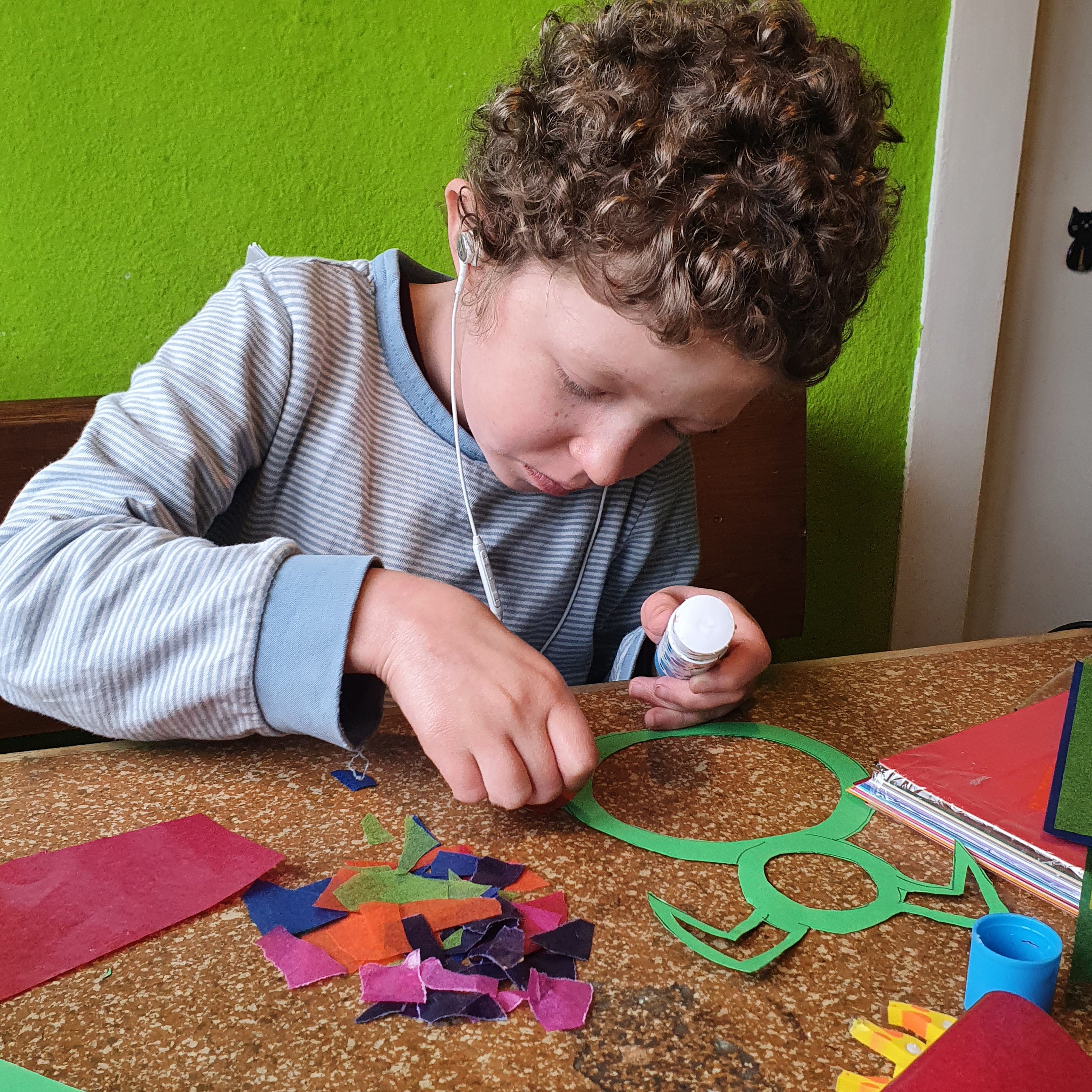 
Ein Kind mit lockigen Haaren sitzt an einem Tisch und bastelt konzentriert. Es trägt ein gestreiftes Langarmshirt und hält eine kleine Klebeflasche in der Hand. Vor ihm liegen bunte Papierschnipsel, rote und violette Bastelbögen sowie ein grüner, ausgeschnittener Rahmen in Hasenform. Im Hintergrund ist eine grüne Wand zu sehen.