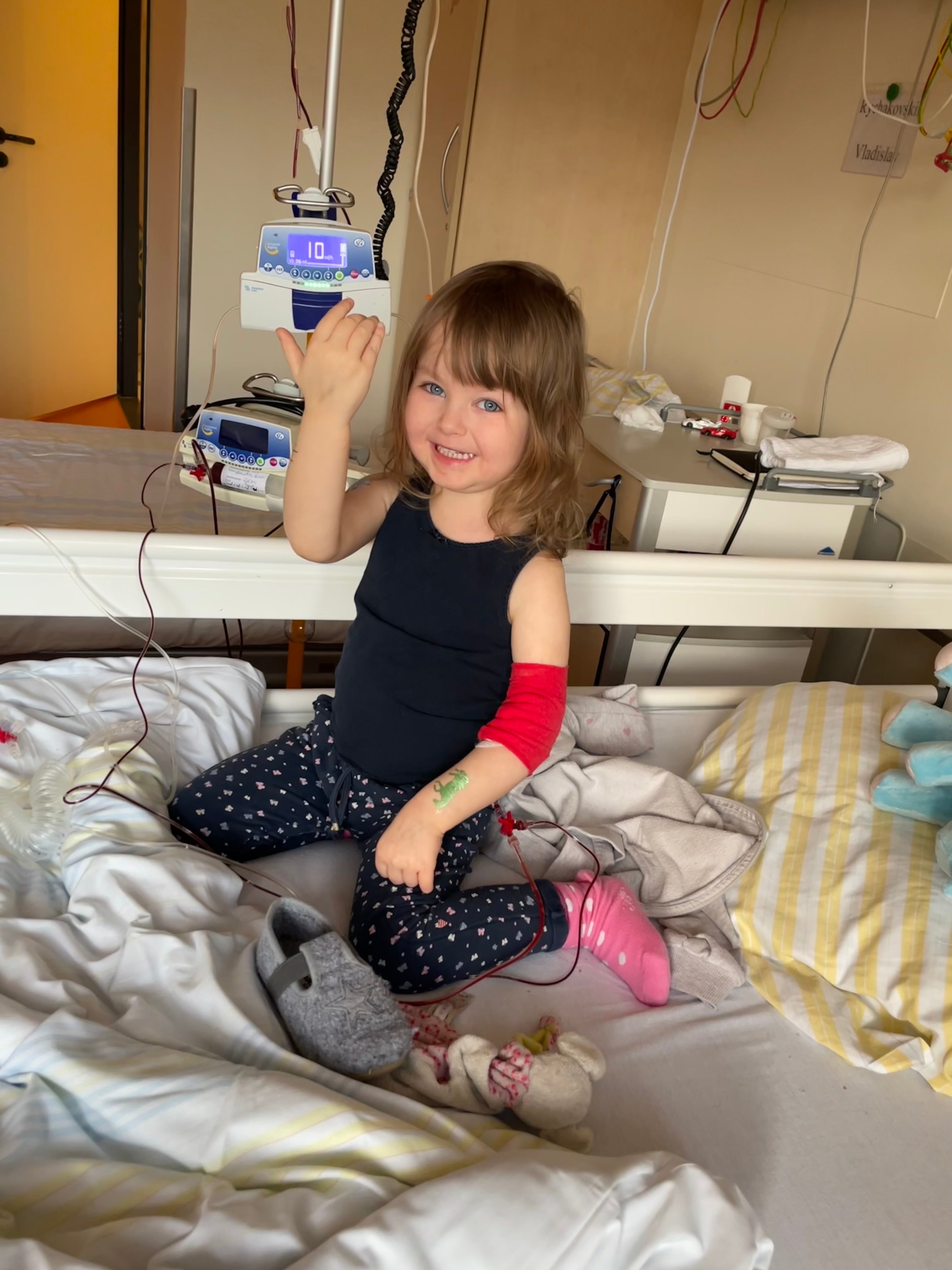 Ein kleines Mädchen sitzt auf einem Krankenhausbett und lächelt in die Kamera. Sie trägt ein dunkles Oberteil, gepunktete Leggings, eine pinke Socke und hat einen roten Verband am rechten Arm. An ihrem Arm ist ein Zugang gelegt, der mit einer Infusionsleitung verbunden ist. Im Hintergrund sind medizinisches Gerät, ein Nachttisch und weitere Gegenstände zu sehen.

