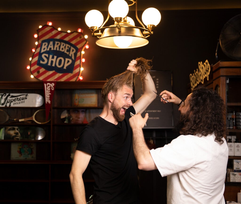 Daniel Korte hält seine langen Haare in einem Zopf hoch. Ein zweiter Mann hält eine Schere vor ihm. 