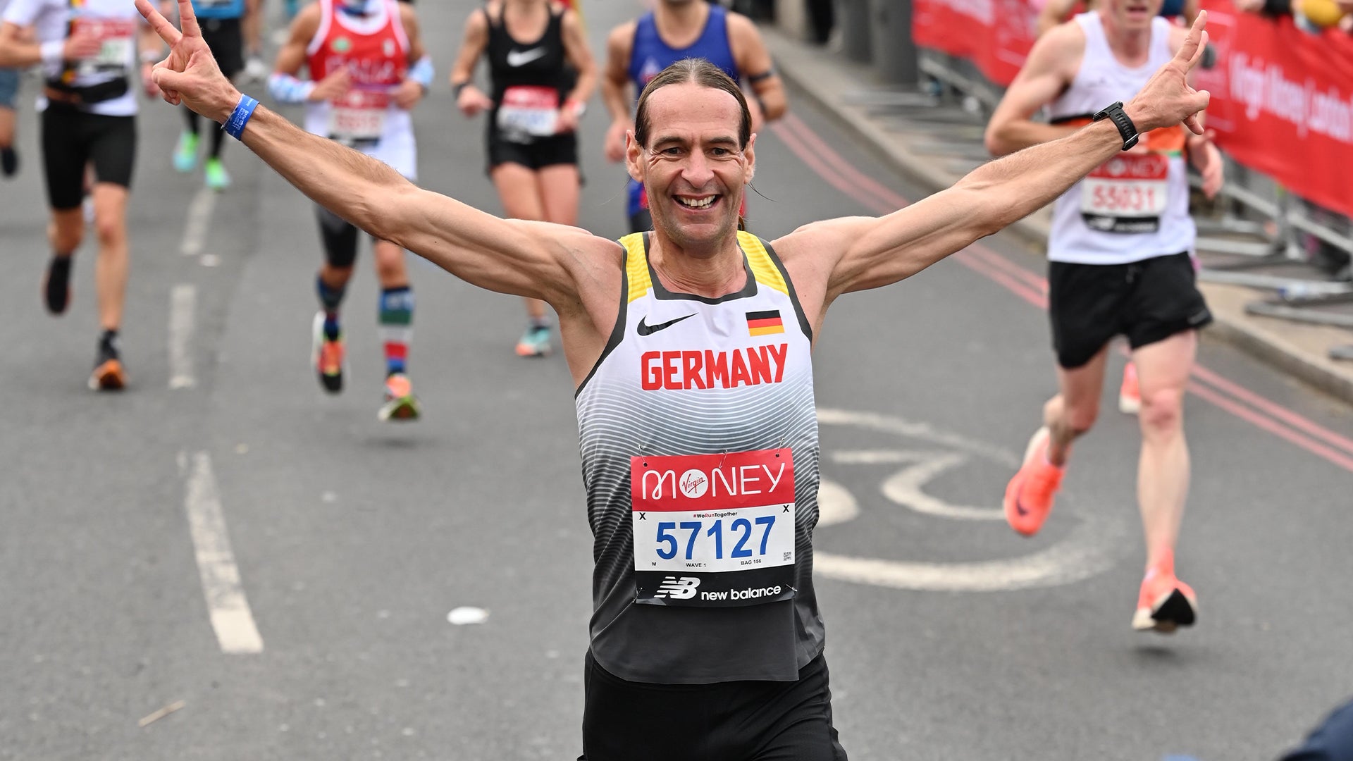 Ein Mann läuft bei einem Straßenmarathon mit ausgestreckten Armen auf die Kamera zu. Er trägt ein Lauftrikot mit der Aufschrift „Germany“, eine Startnummer mit der Ziffer 57127 und ein breites Lächeln im Gesicht. Hinter ihm sind weitere Läuferinnen und Läufer auf der abgesperrten Straße zu sehen.
