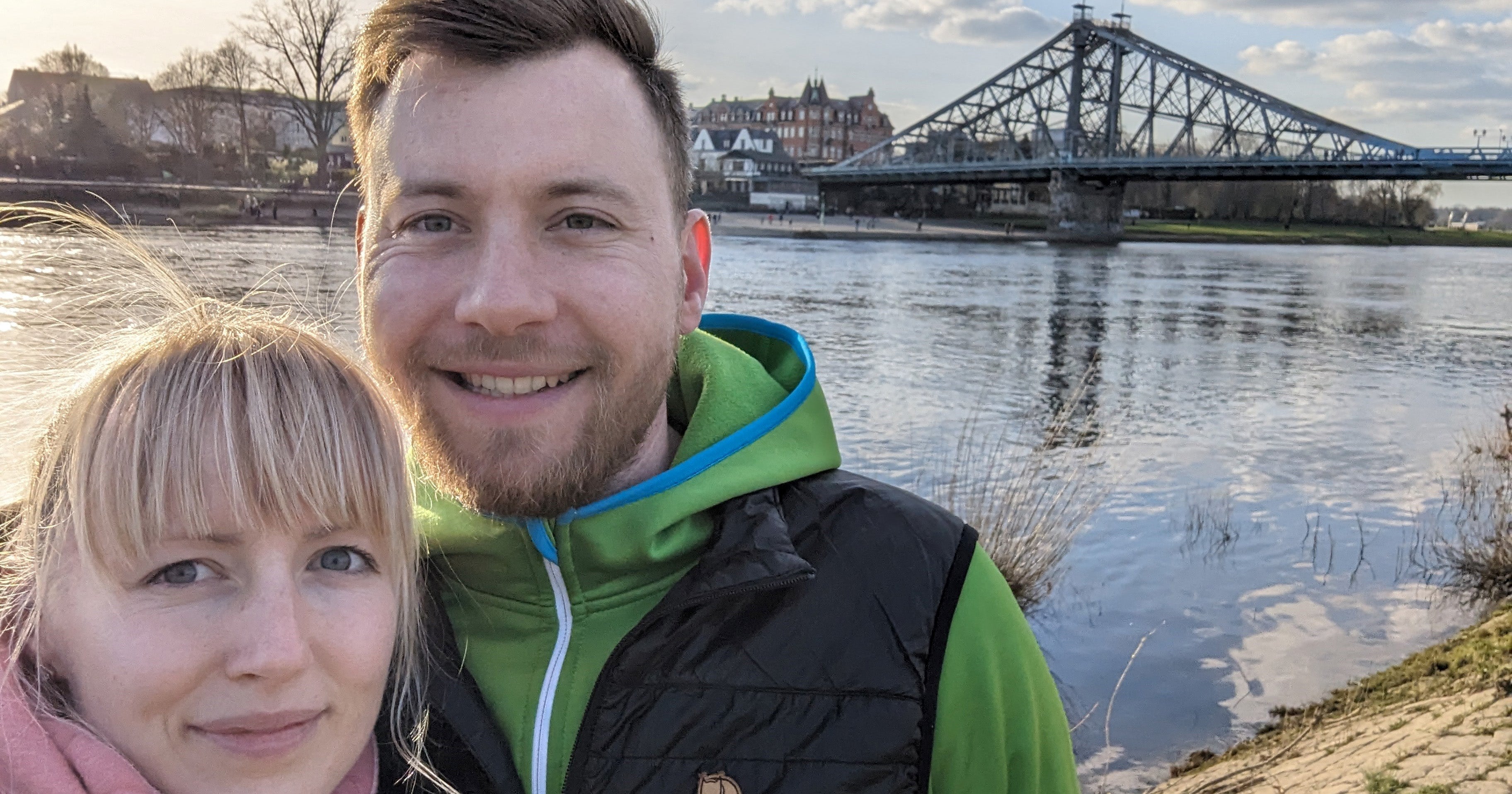 Eine Frau und ein Mann lächeln in die Kamera. Sie stehen am Flußufer und im Hintergrund ist eine Brücke zu sehen.