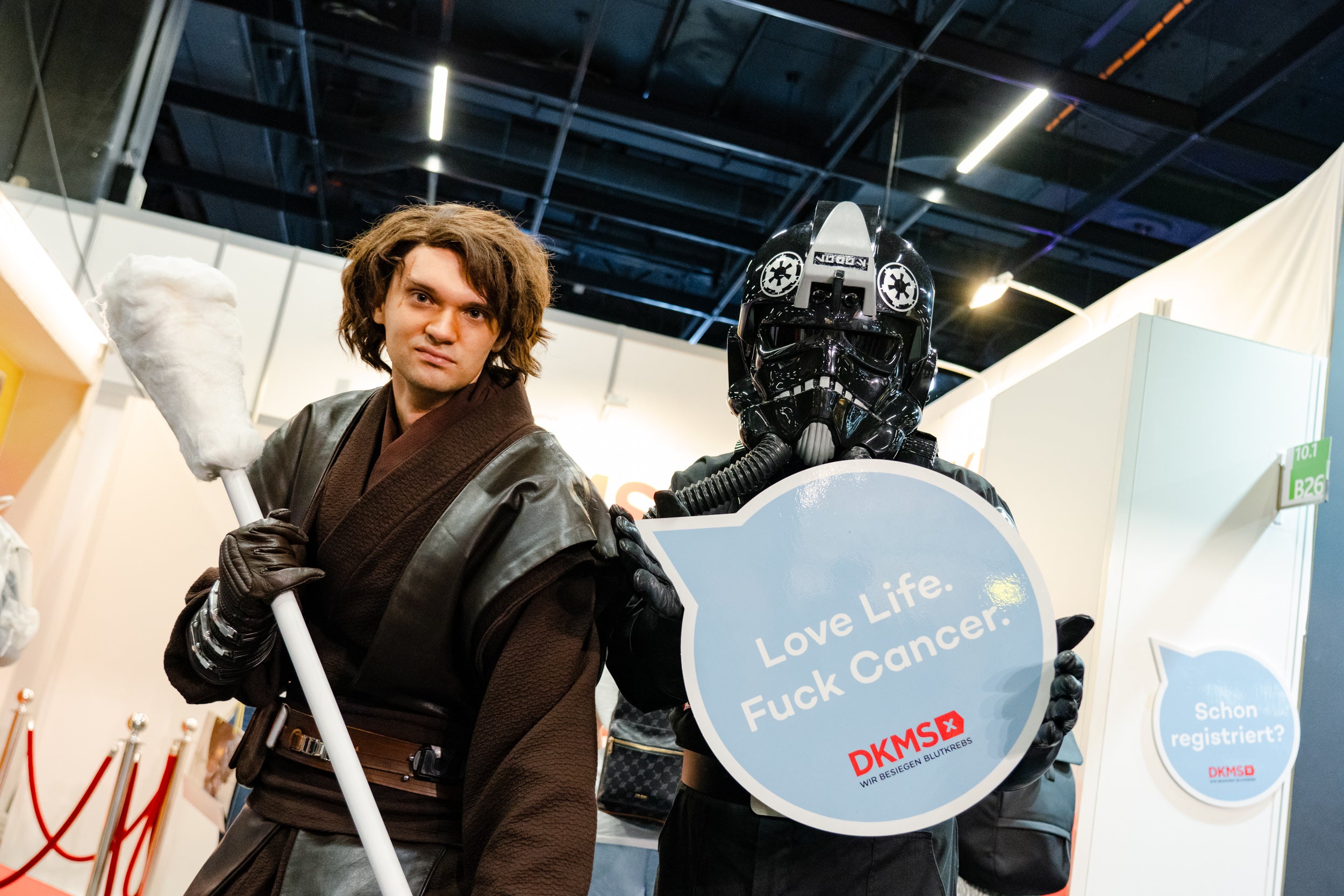 Zwei Personen in Star-Wars-Kostümen stehen vor einem Messestand. Die linke Figur trägt ein braunes Jedi-Kostüm und hält ein übergroßes Wattestäbchen. Die rechte Figur trägt eine schwarze Pilotenrüstung und hält ein Schild mit der Aufschrift: „Love Life. Fuck Cancer. – DKMS“. Im Hintergrund sind Messestände, weiße Trennwände und weitere Sprechblasen-Schilder zu sehen.
