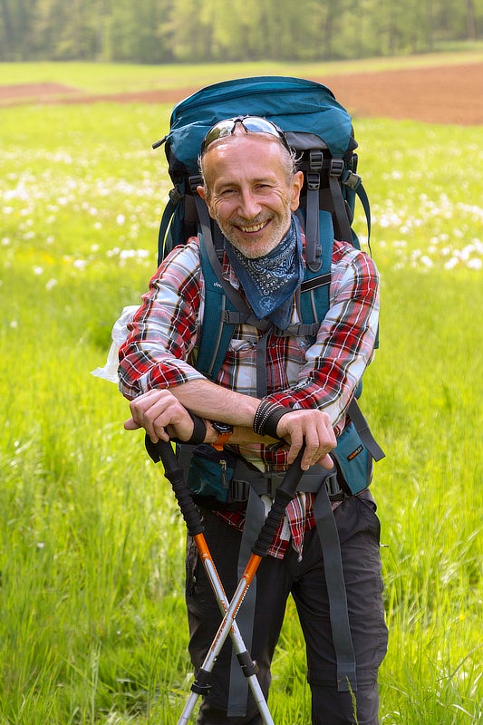Fuat Gören mit einem Rucksack. Im Hintergrund ist eine Blumenwiese.
