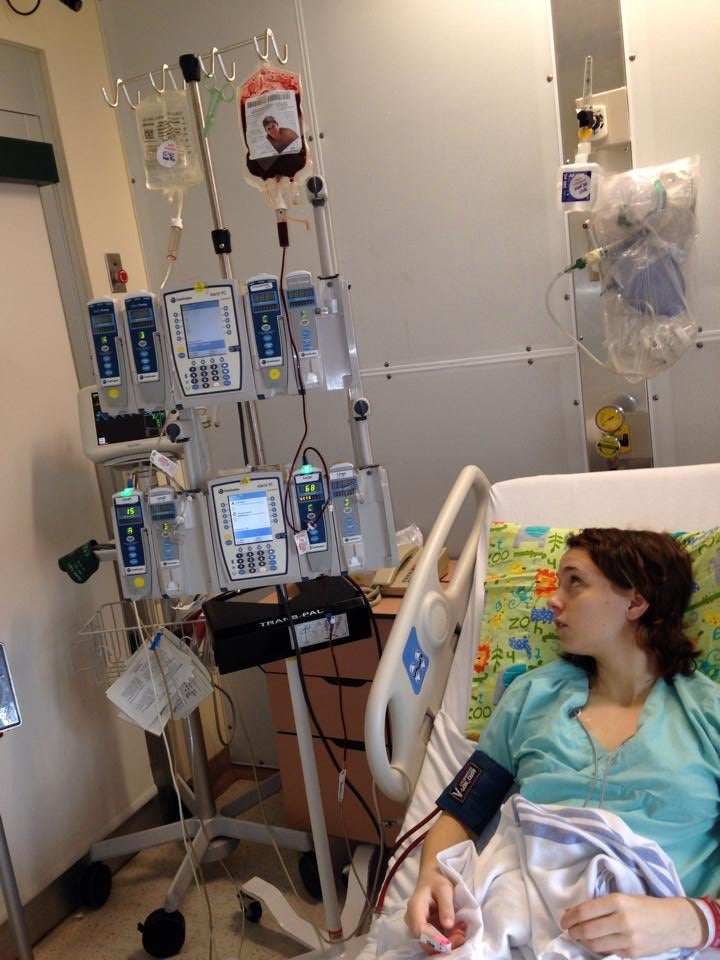 Eine junge Patientin liegt in einem Krankenhausbett und blickt nach oben zu mehreren Infusionspumpen. An einem Infusionsständer hängen mehrere Beutel mit Flüssigkeiten und eine Bluttransfusion. Die Geräte sind an Monitore und Schläuche angeschlossen. Die Patientin trägt ein Krankenhaushemd, ein Blutdruckmessgerät am Arm und liegt unter einer Decke mit bunter Bettwäsche. Die Umgebung ist ein klinisch ausgestattetes Krankenzimmer.