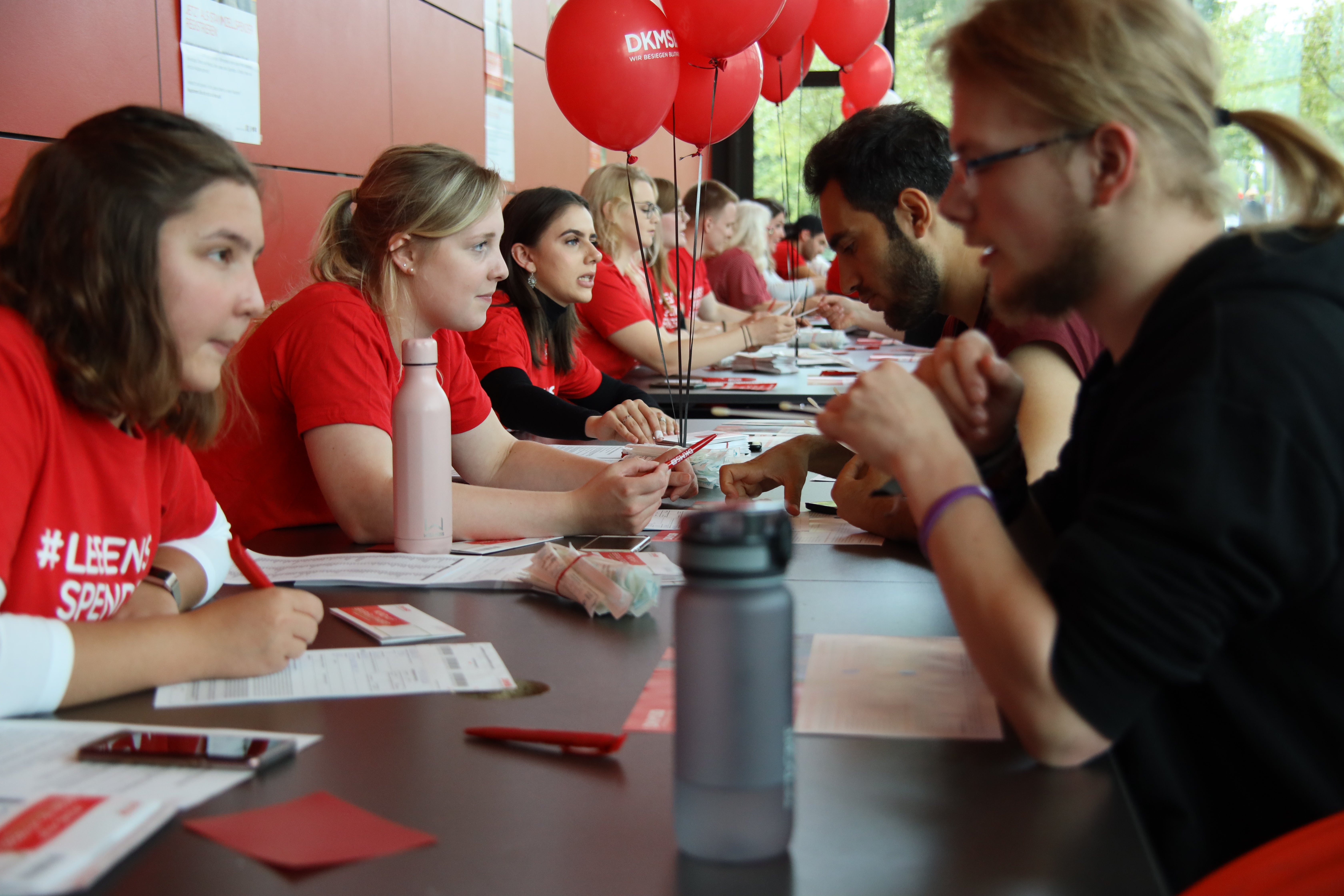 Mehrere junge Menschen in roten DKMS-T-Shirts sitzen an einem langen Tisch und beraten interessierte Personen gegenüber. Auf dem Tisch liegen Infomaterialien und Wasserflaschen. Rote DKMS-Luftballons sind im Hintergrund zu sehen