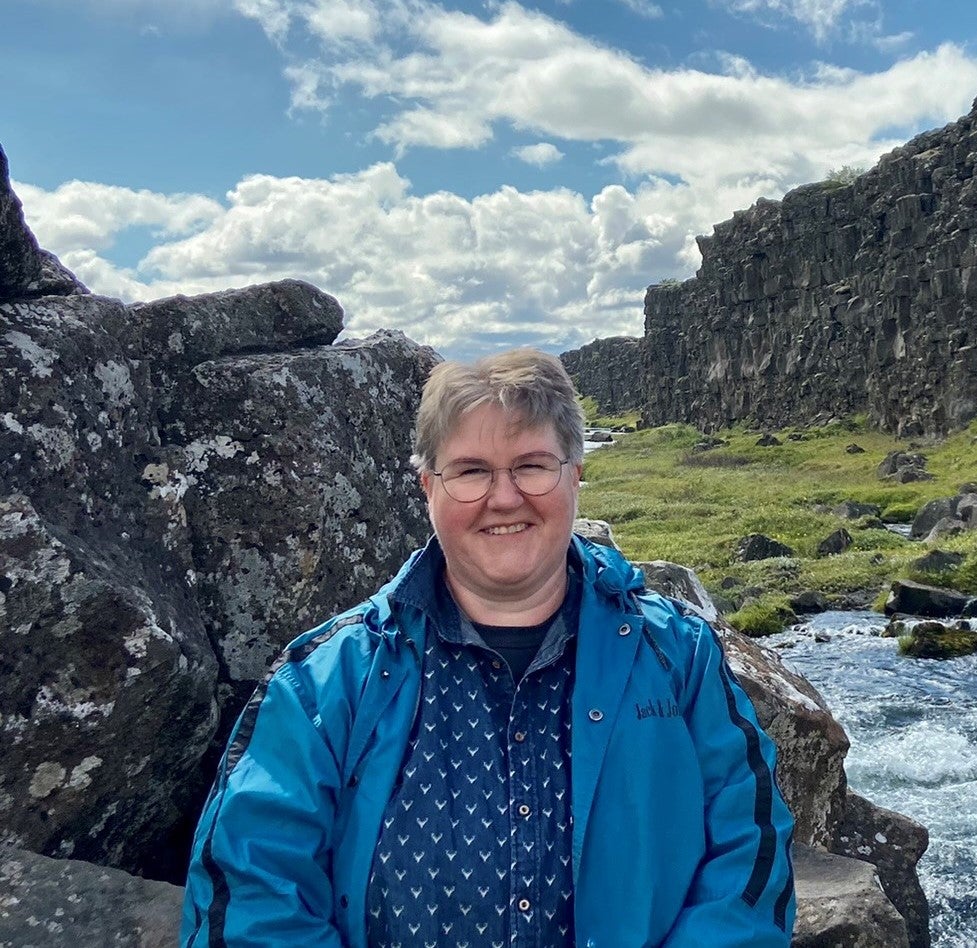 eine Frau sitzt auf einem Stein, im Hintergrund sieht man einen Fluss und Berge