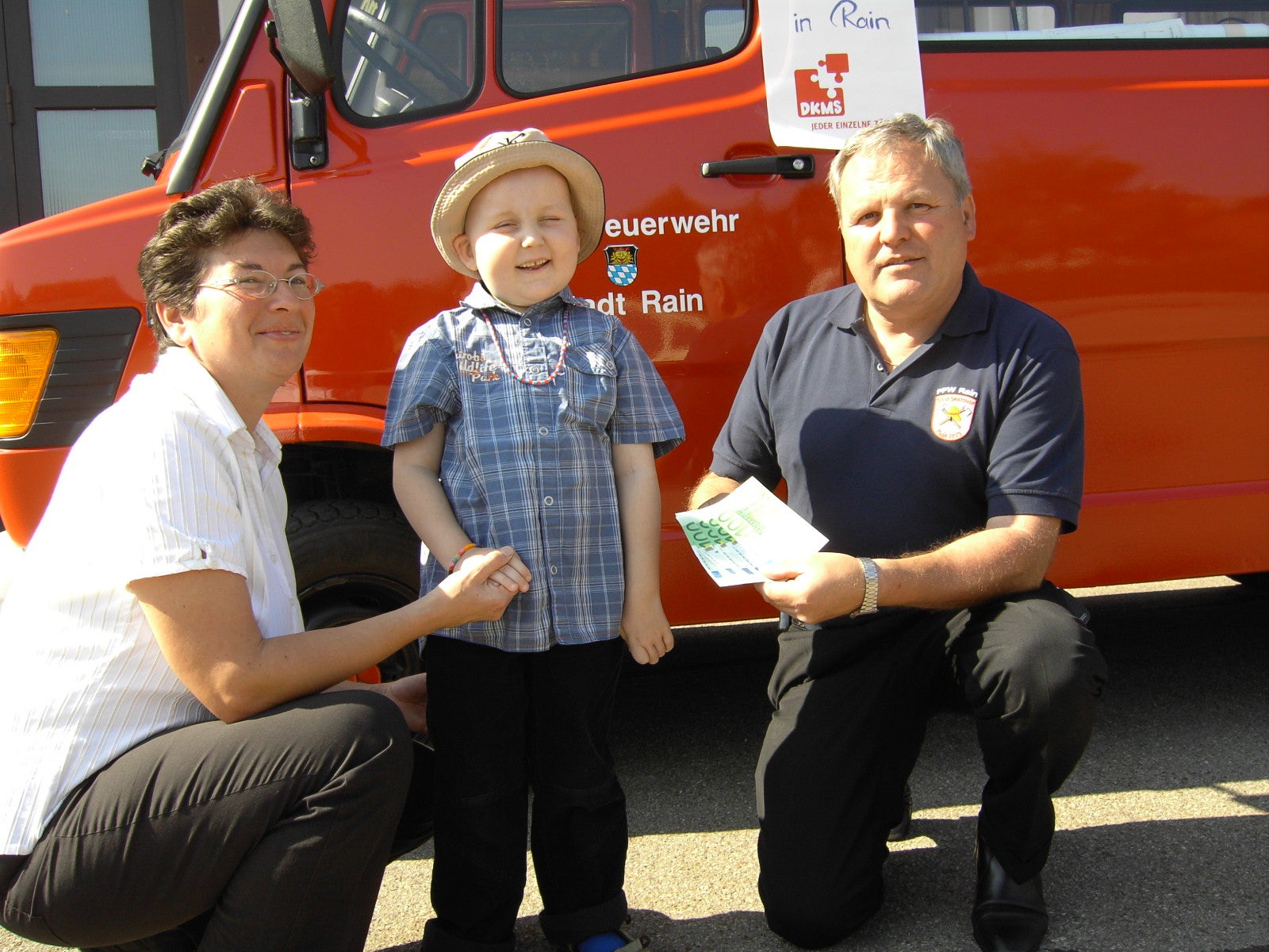 Brigitte Lehenberger mit einem kleinen Kind und einem Mann, kniend vor einem Feuerwehrauto