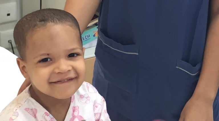 Ein Kind lächelt in die Kamera, rechts daneben steht eine Person in einem Kasack