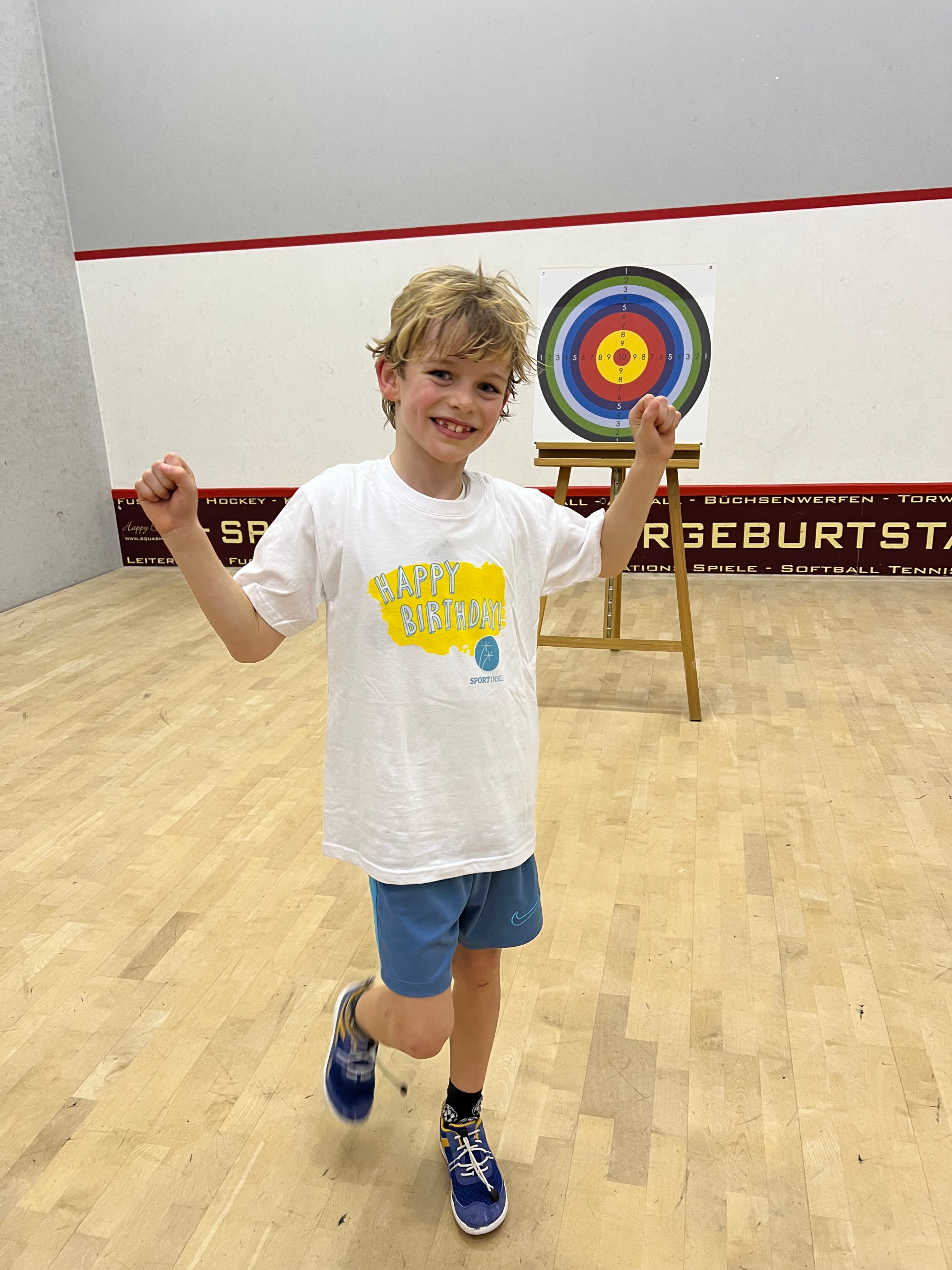 Ein Junge in einem weißen T-Shirt und blauen Shorts hat die Arme nach oben gehoben und lächelt.