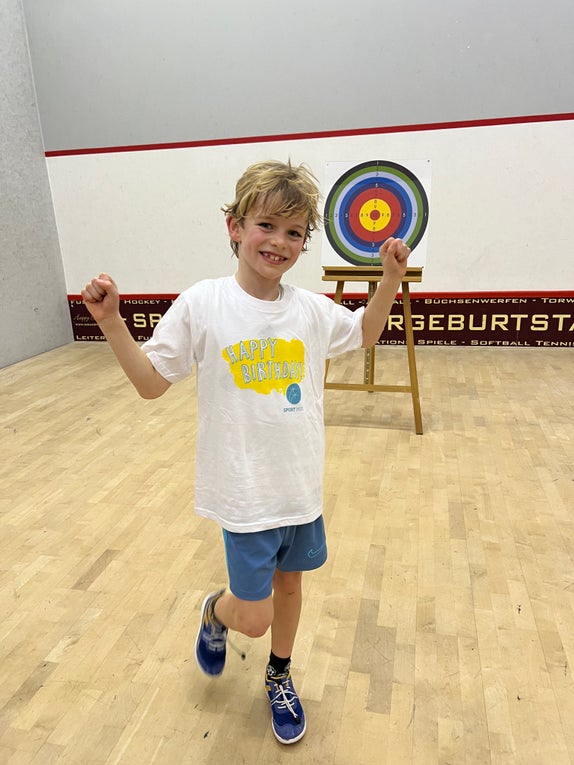 Ein Junge in einem weißen T-Shirt und blauen Shorts hat die Arme nach oben gehoben und lächelt.