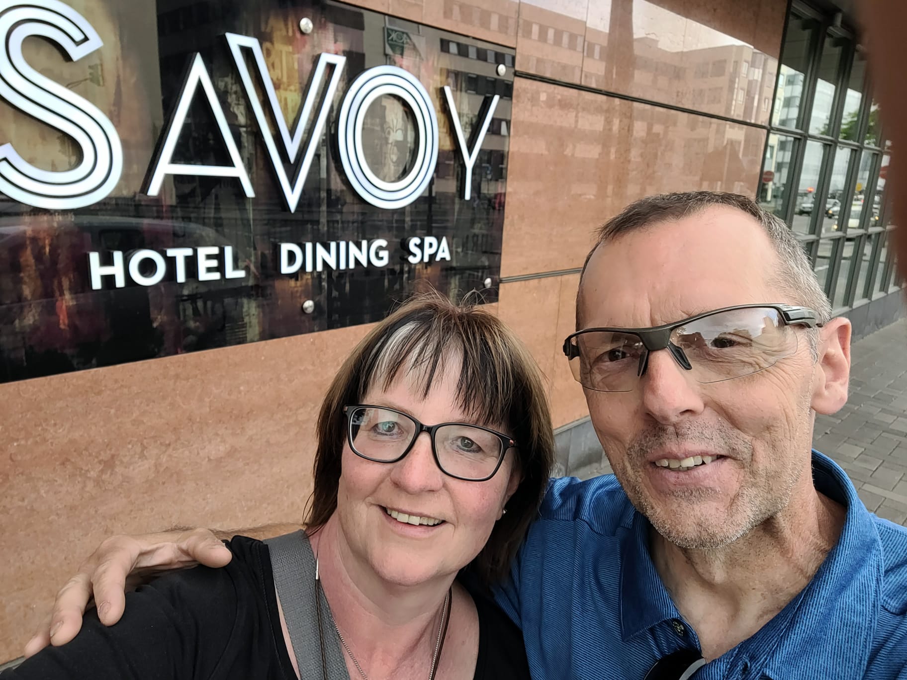 Ein Mann und eine Frau stehen vor einer Fassade mit der Aufschrift „Savoy Hotel Dining Spa“ und machen ein Selfie. Beide tragen Brillen, der Mann ein blaues Hemd und eine Sportbrille, die Frau ein schwarzes Oberteil. Die Hauswand besteht aus braunem Stein. Im Hintergrund spiegeln sich Gebäude in den Fenstern.
