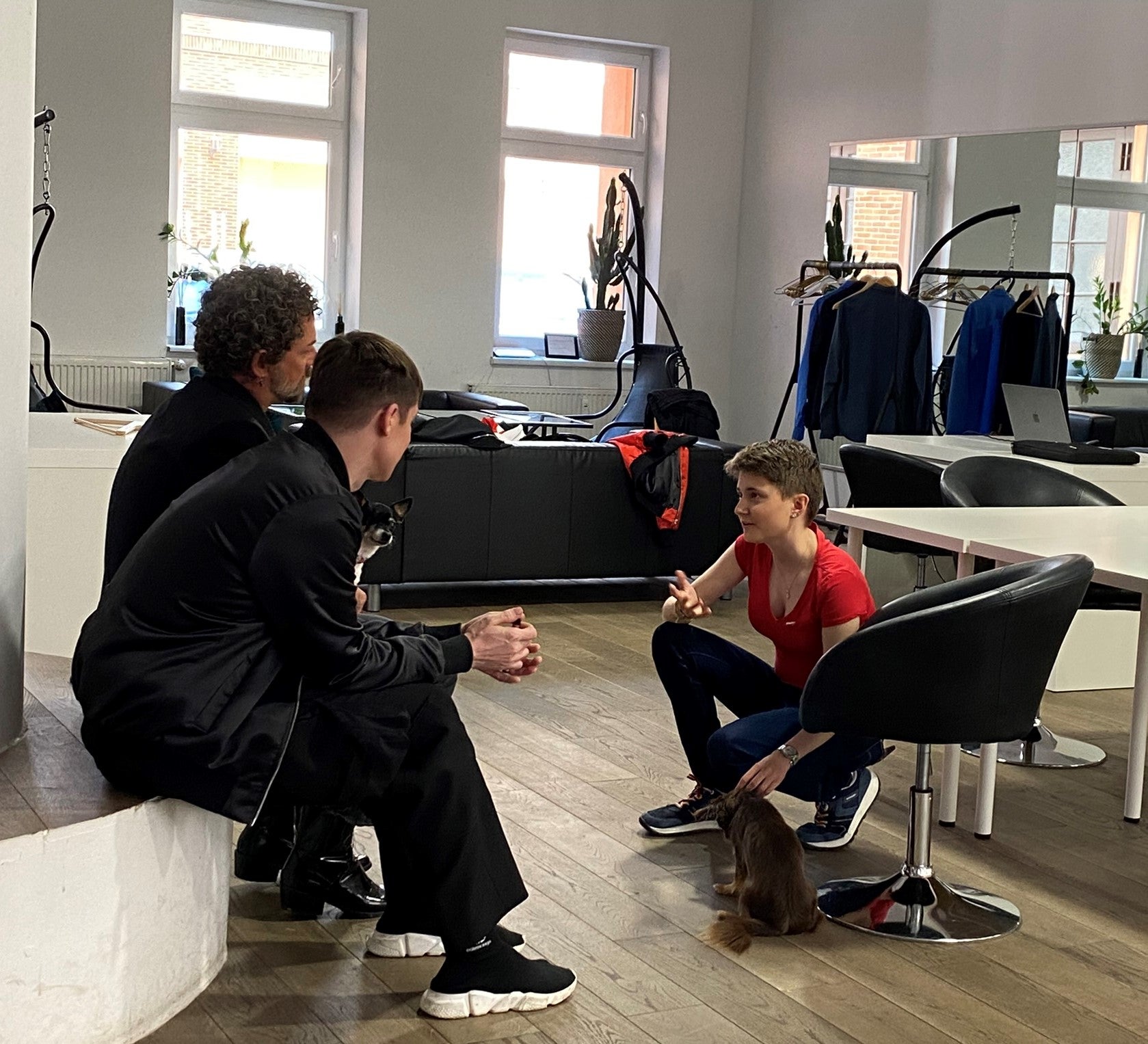 Zwei Männer sitzen auf einer niedrigen Mauer, eine Frau in rotem T-Shirt und Jeans hockt ihnen gegenüber auf dem Boden. Die drei Personen befinden sich in einem hellen Raum mit großen Fenstern, Friseurstühlen und Kleiderständern im Hintergrund. Auf dem Boden liegt ein brauner Perückenzopf.
